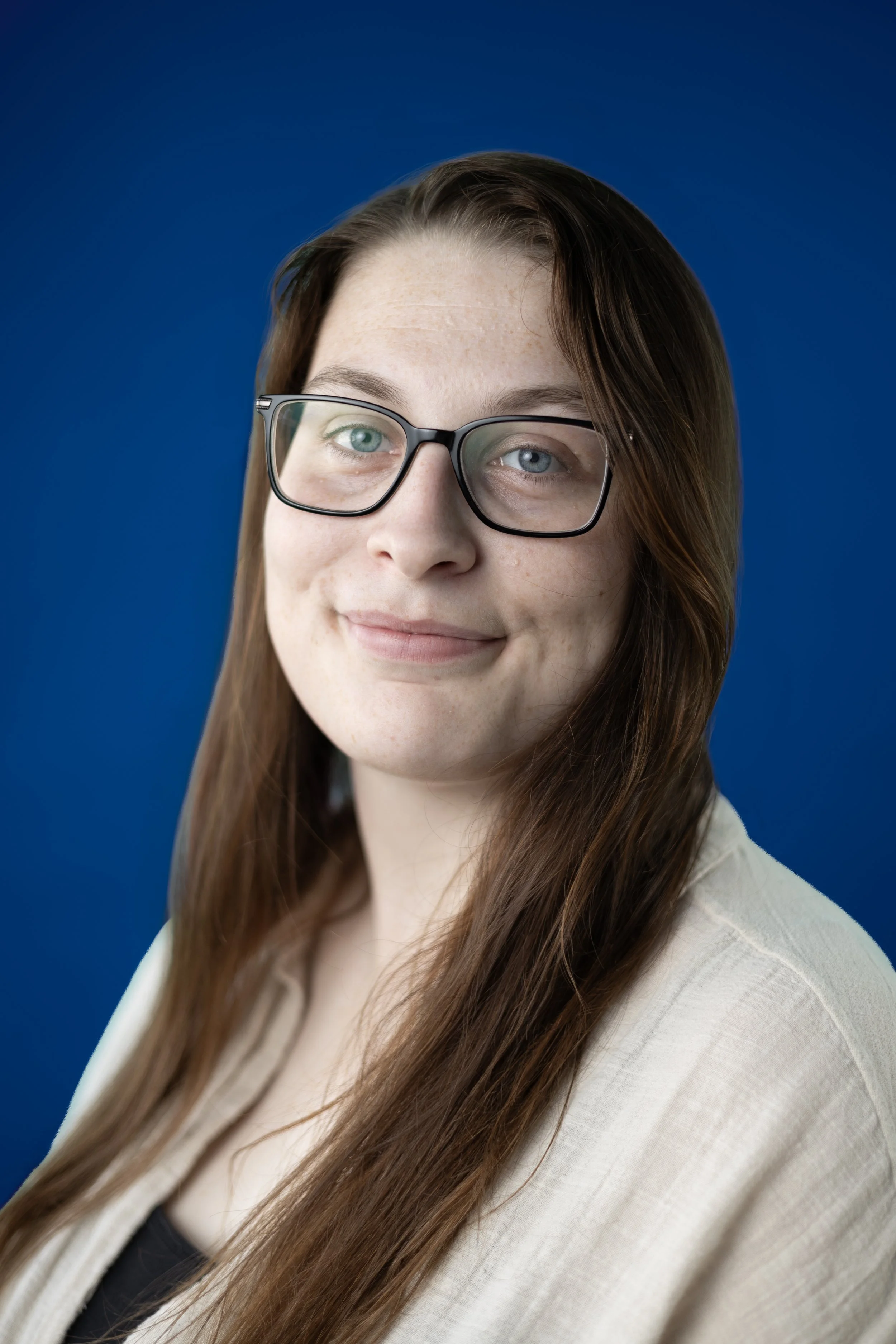 A young woman with long brown hair, wearing glasses and a beige blazer, is smiling and looking slightly to the side against a blue background.