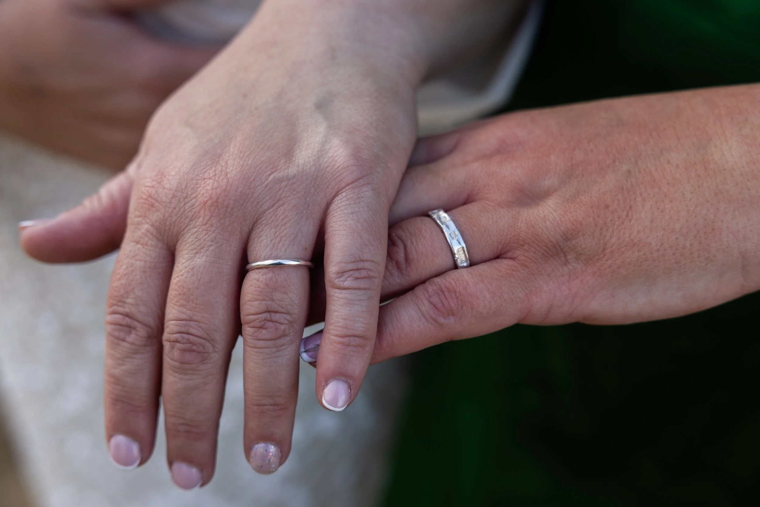 Close-up of two hands clasped together, showing wedding rings on the ring fingers. One hand has a plain silver band, and the other has a silver band with small embedded stones. The background is blurred.