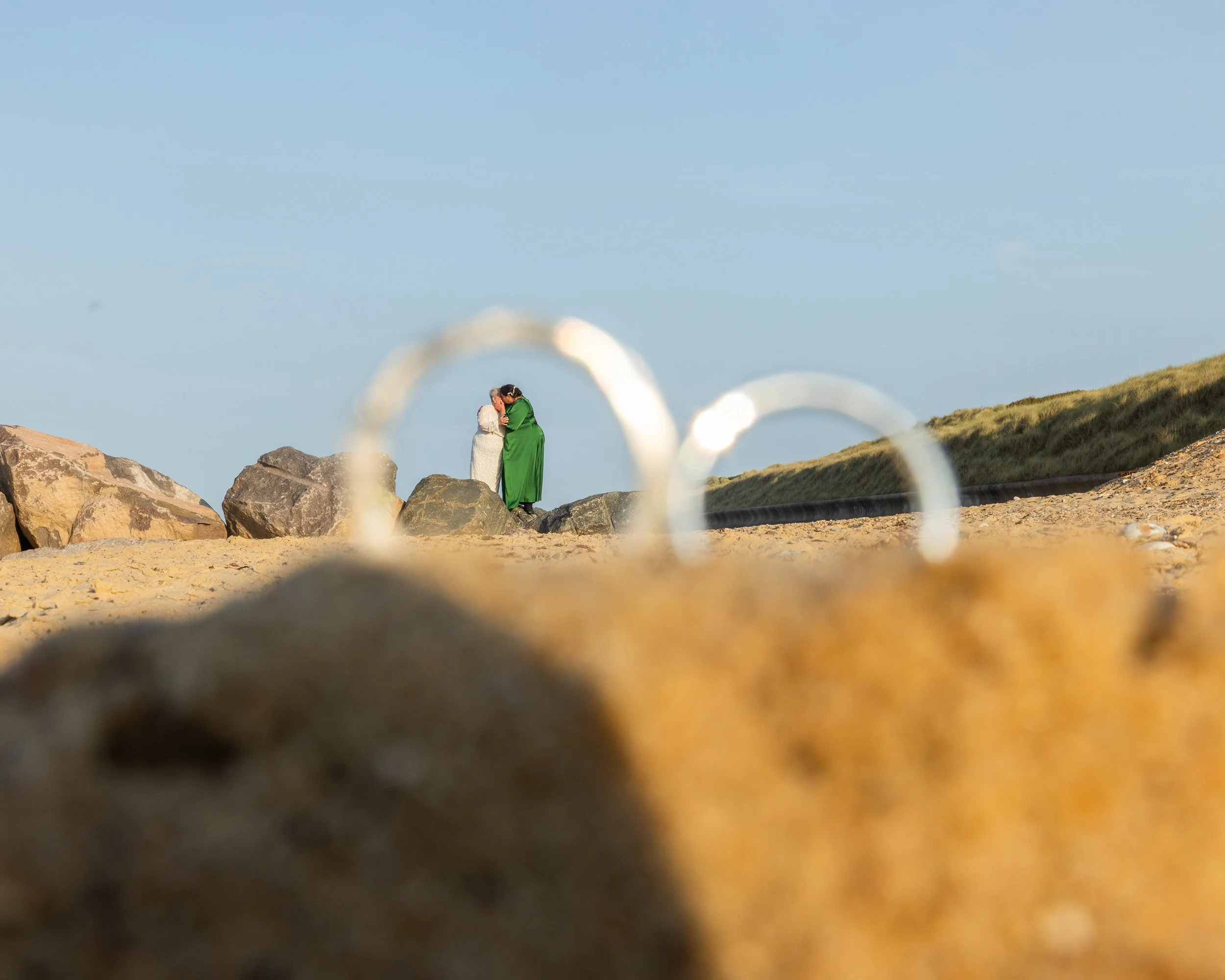 Two people, likely a bride and groom, standing on rocks near the water, embracing each other, with a scenic landscape of grasses and hills in the background, viewed through a blurred foreground.
