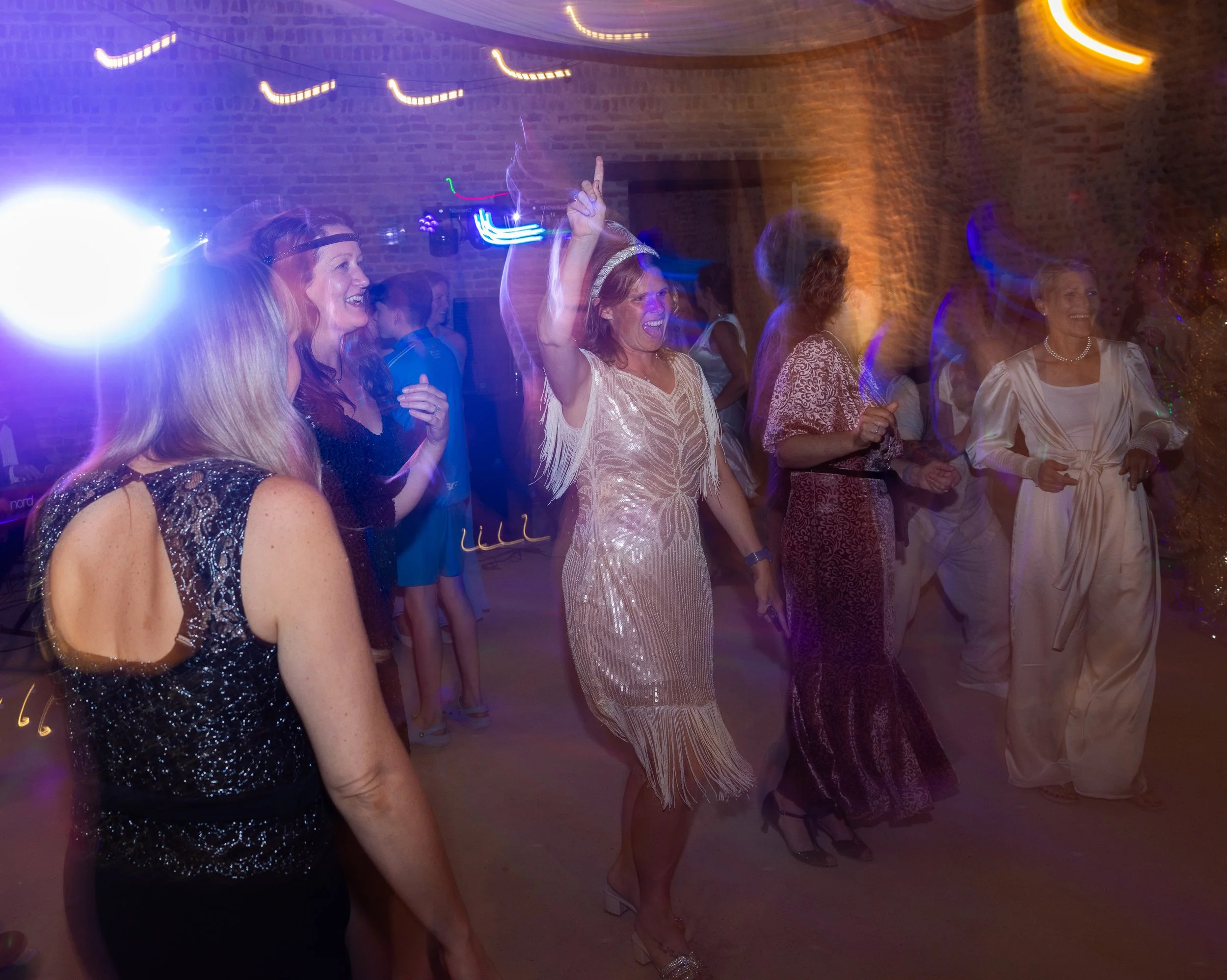 People dancing and enjoying themselves at a nighttime party or celebration, with colorful lights and a brick wall in the background.