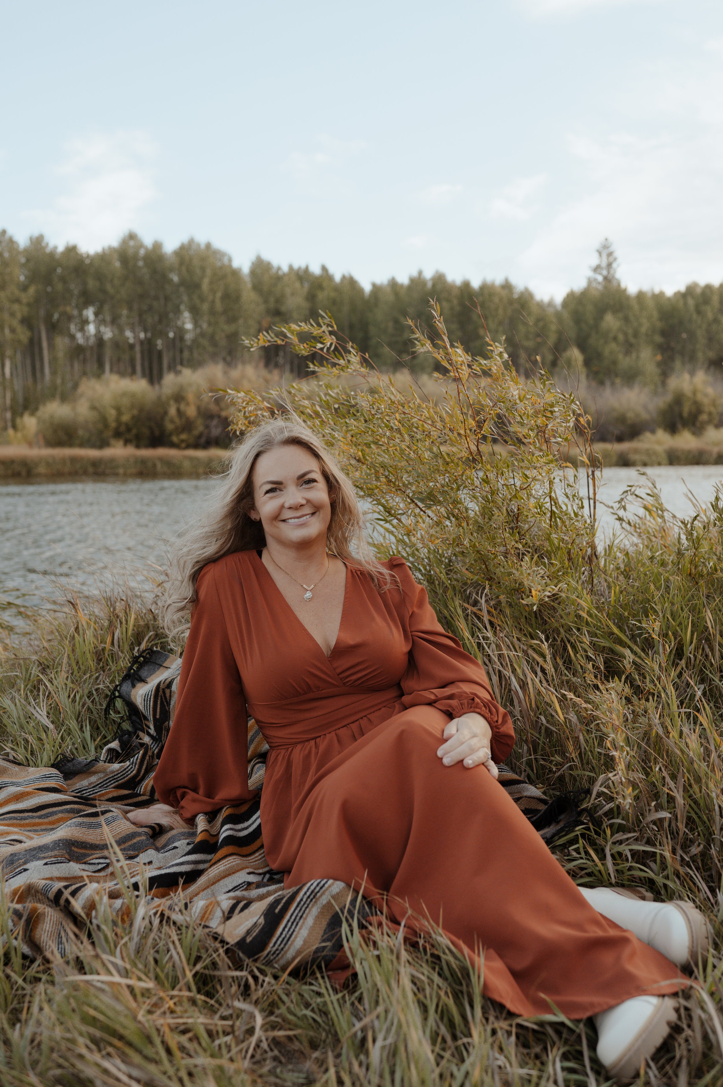 A woman in a rust-colored dress sitting on a striped blanket beside a body of water, with trees and a cloudy sky in the background.