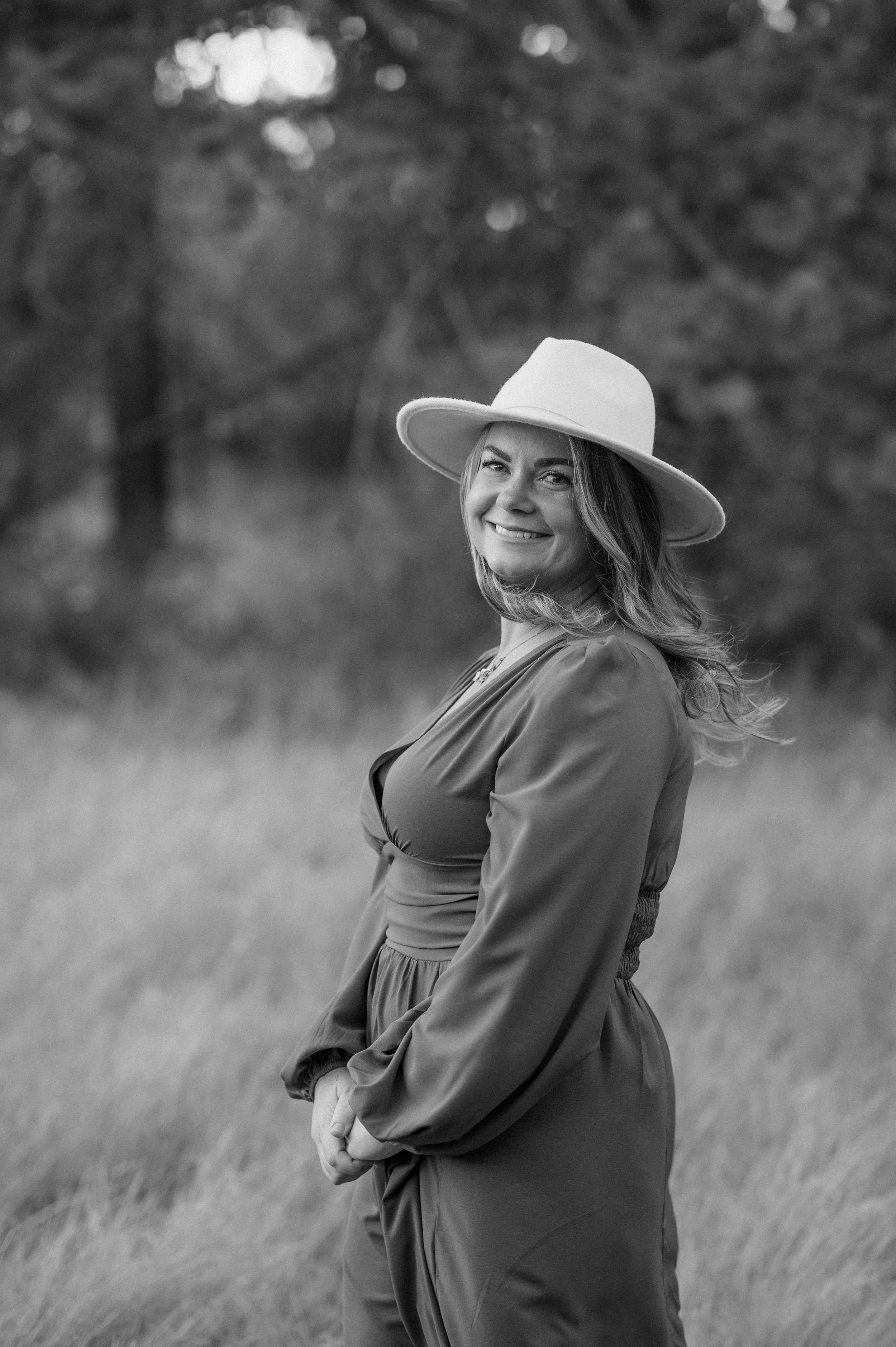 A woman smiling and wearing a wide-brimmed hat and a long-sleeved dress outdoors, in a natural setting with blurred trees and grass