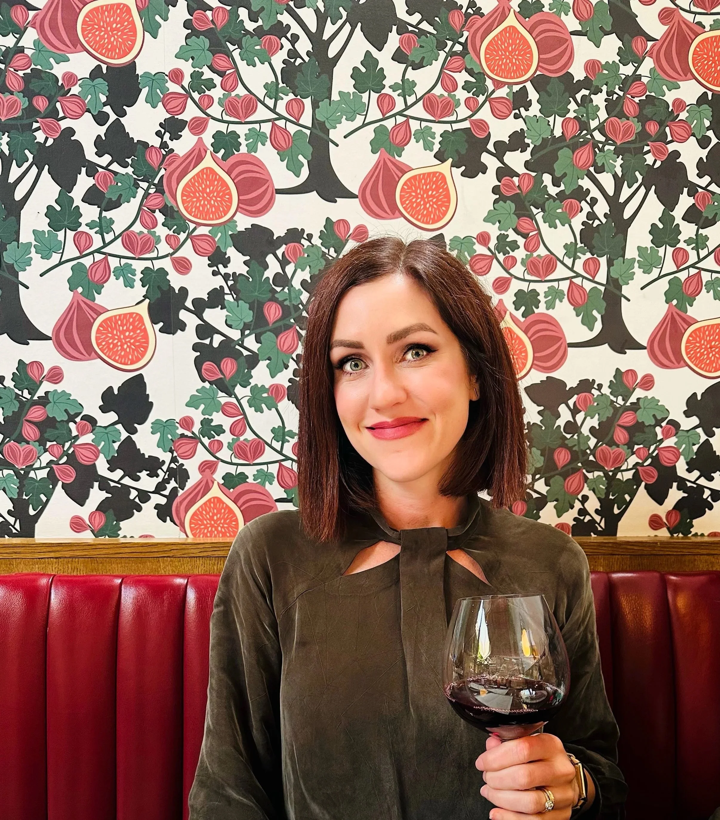 A woman with brown hair and green eyes holding a glass of red wine in a restaurant with a red leather booth seat and a decorative floral wallpaper background.
