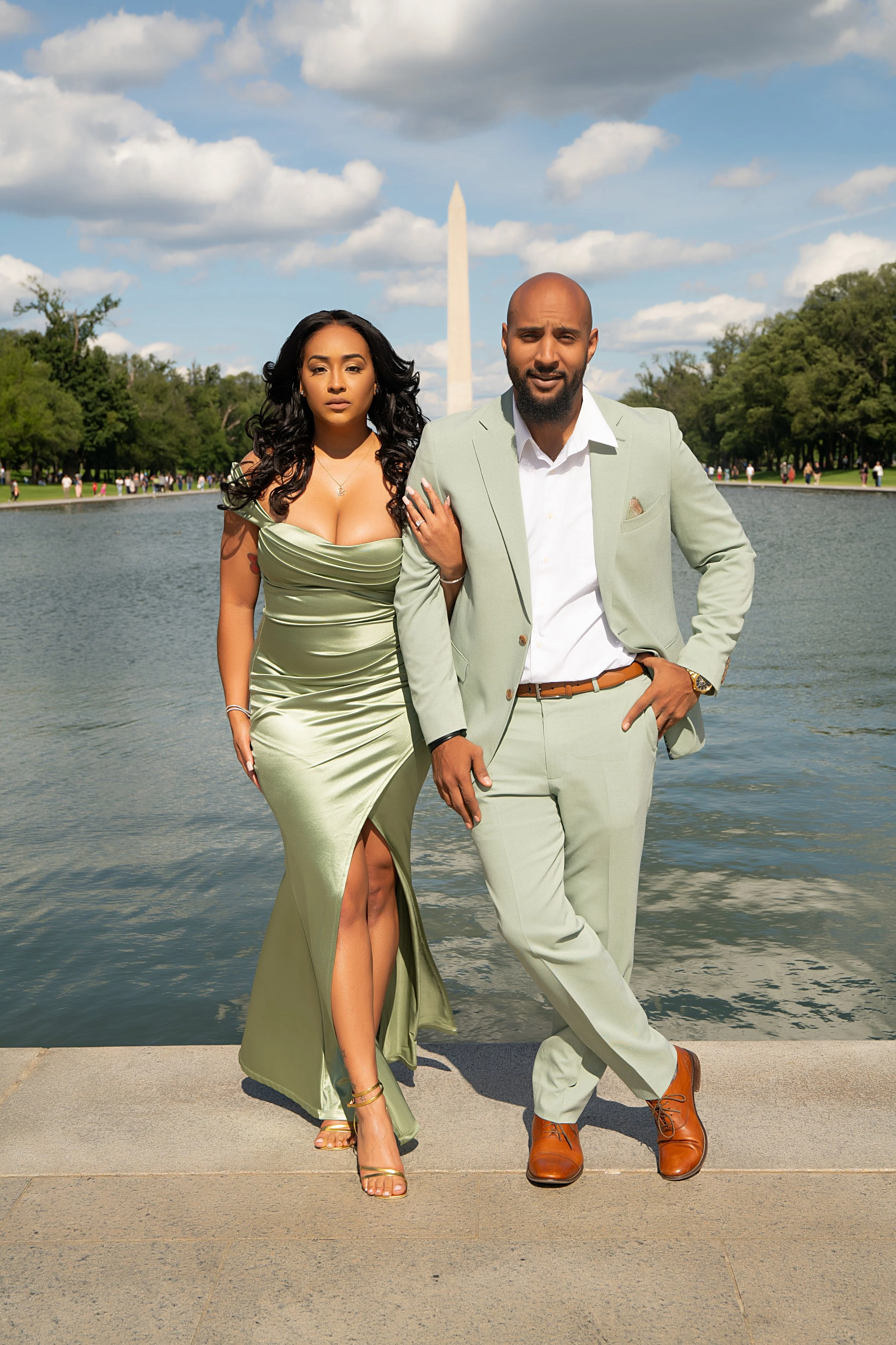 A fashionable man and woman standing by a water feature at the National Mall in Washington, D.C., with the Washington Monument in the background on a sunny day with partly cloudy skies.