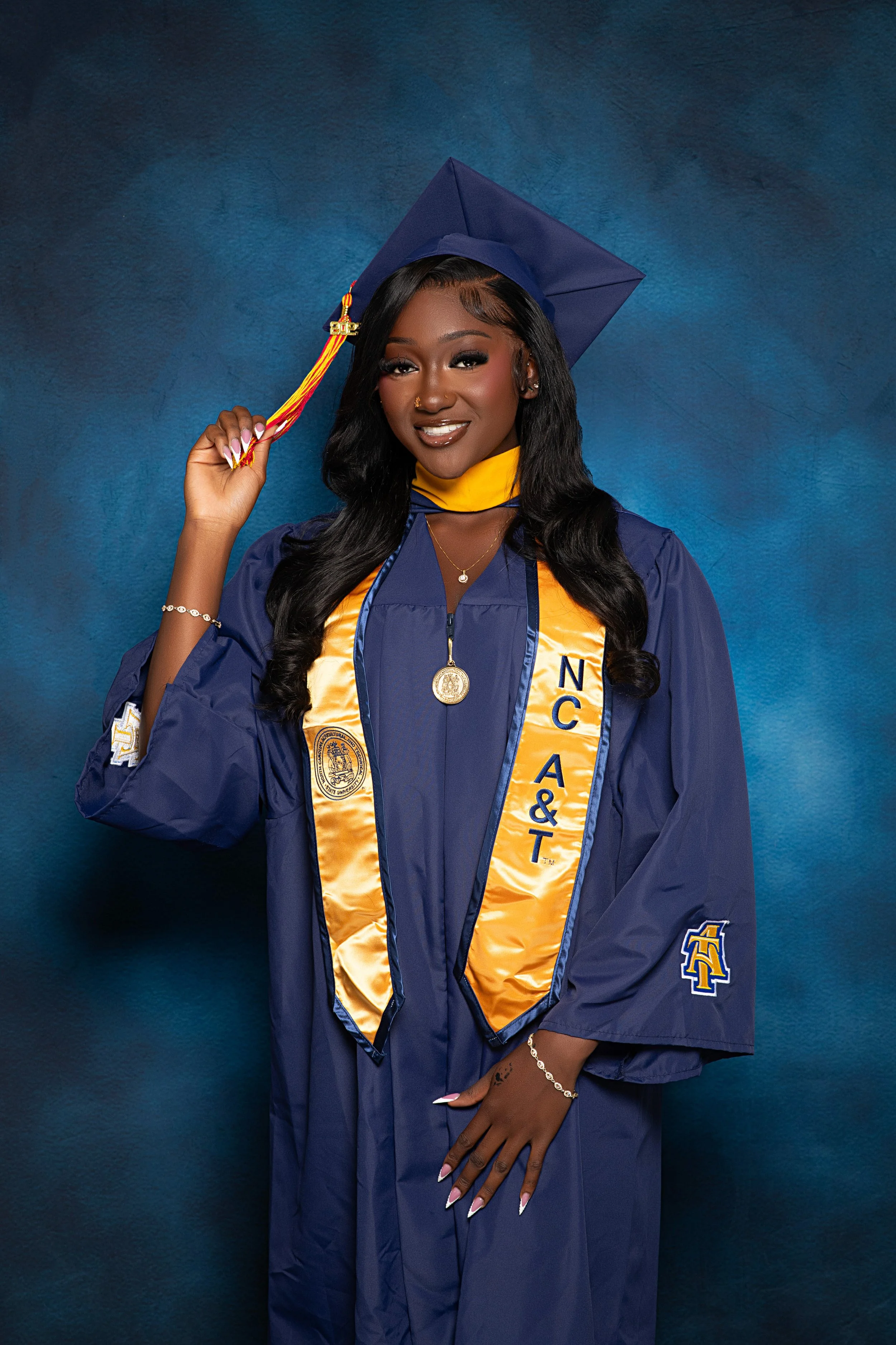 A young woman in a blue graduation gown and cap with a yellow sash, featuring the letters 'NCA&T,' standing against a dark blue background, smiling while holding the tassel of her cap.