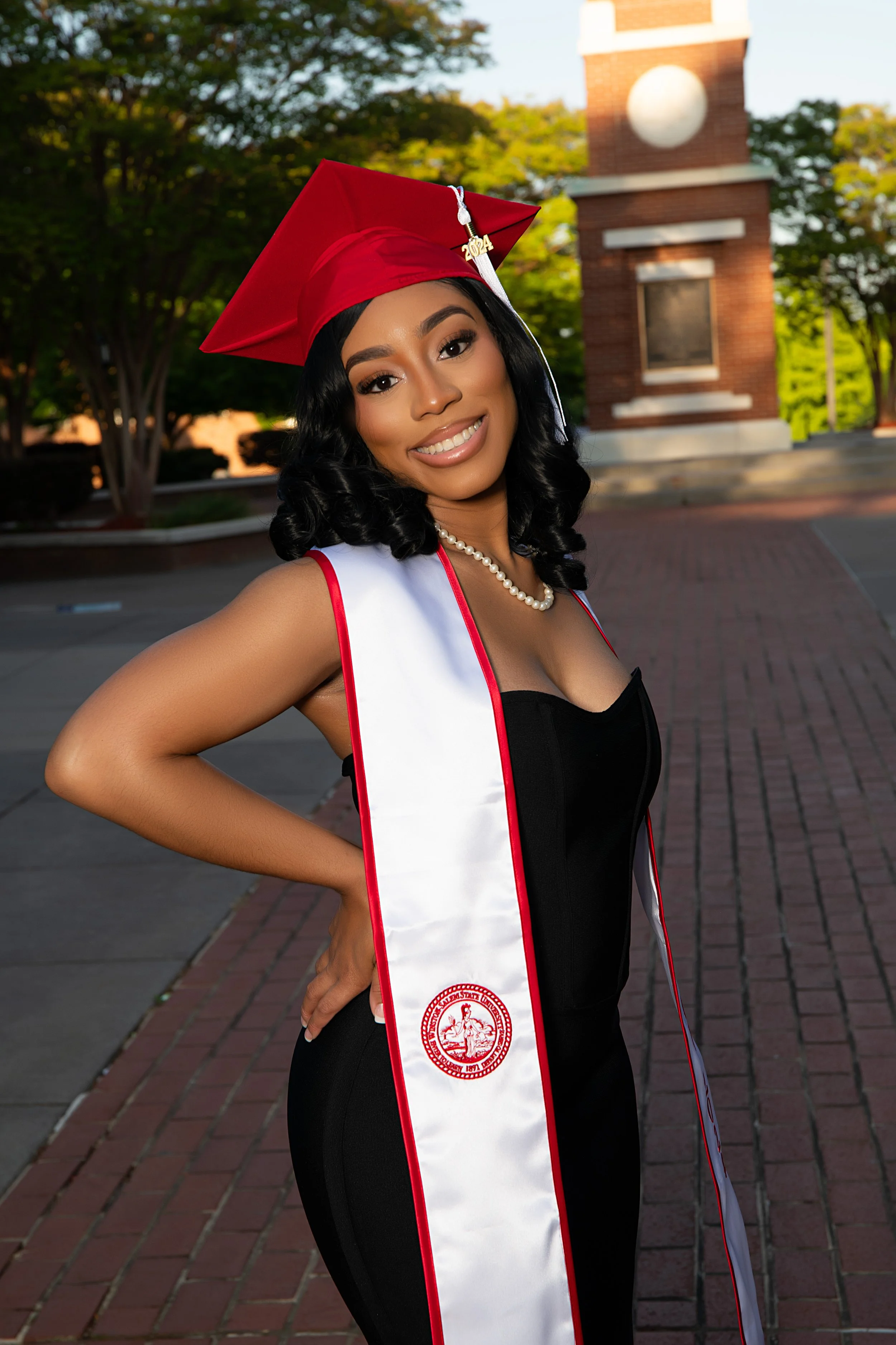 A young woman smiling, dressed in black and red graduation gown and cap, with a white and red sash. She is wearing a pearl necklace and stands outdoors in front of a brick clock tower, with trees in the background.