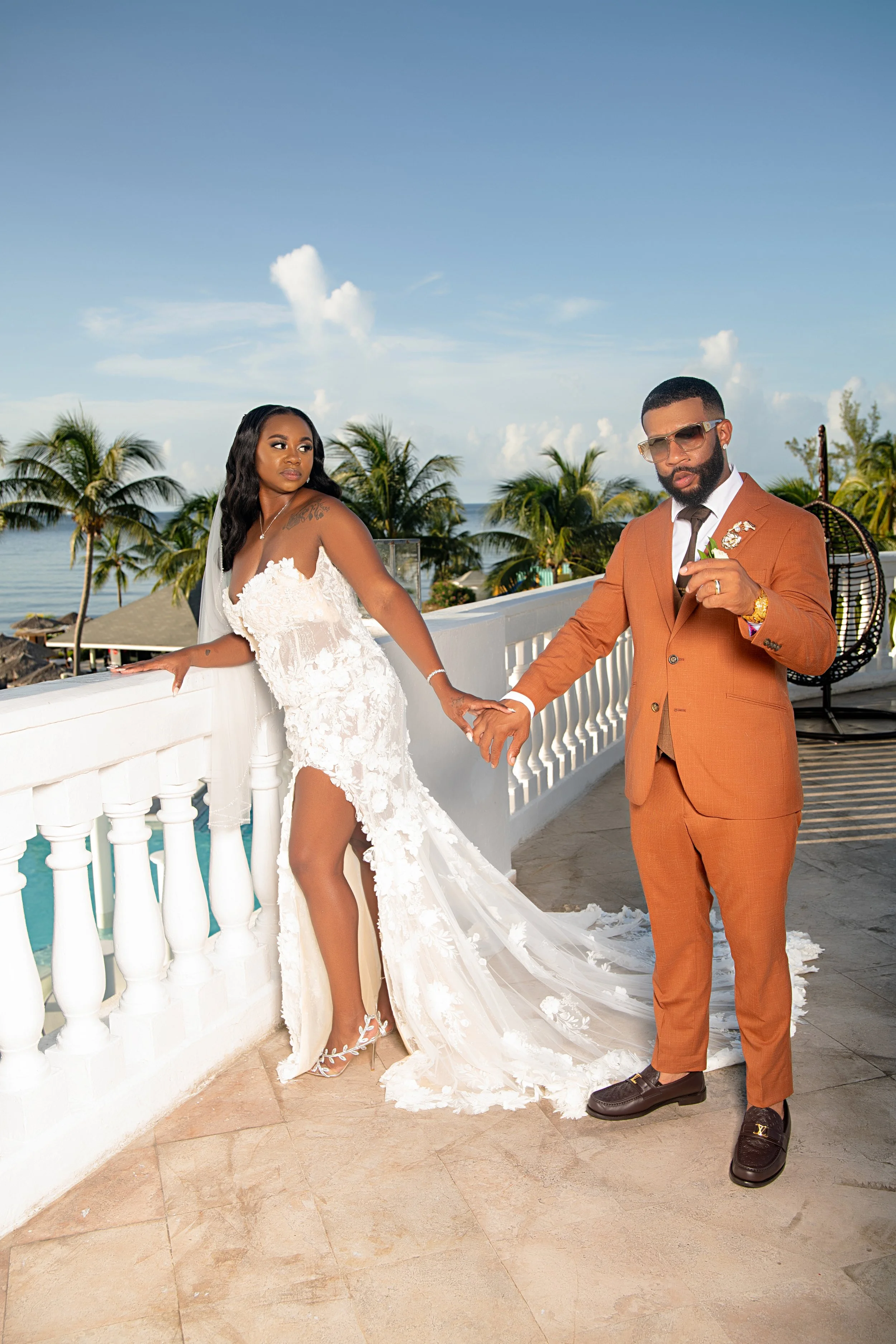 A couple dressed in wedding attire holding hands on a balcony with tropical trees and the ocean in the background, under a blue sky with clouds.