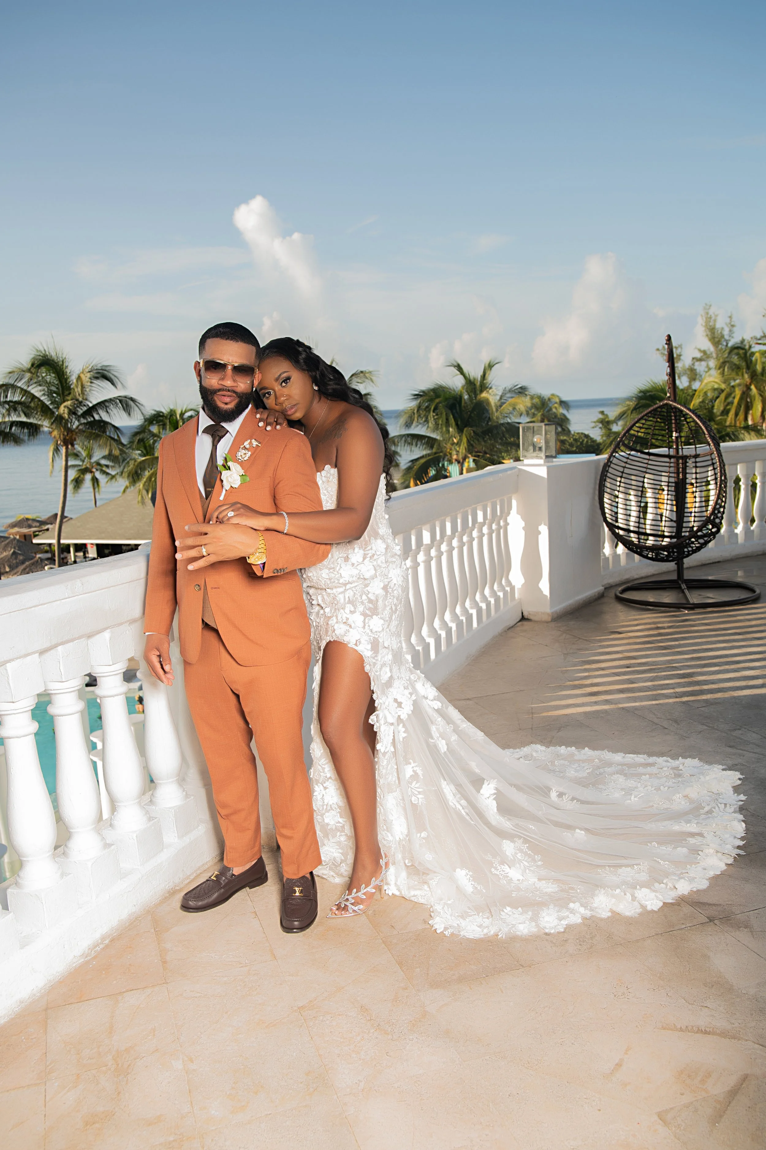 A couple dressed in wedding attire standing on a balcony overlooking a tropical beach with palm trees and the ocean. The groom wears an orange suit and sunglasses, and the bride wears a strapless wedding gown with a long train and lace details.