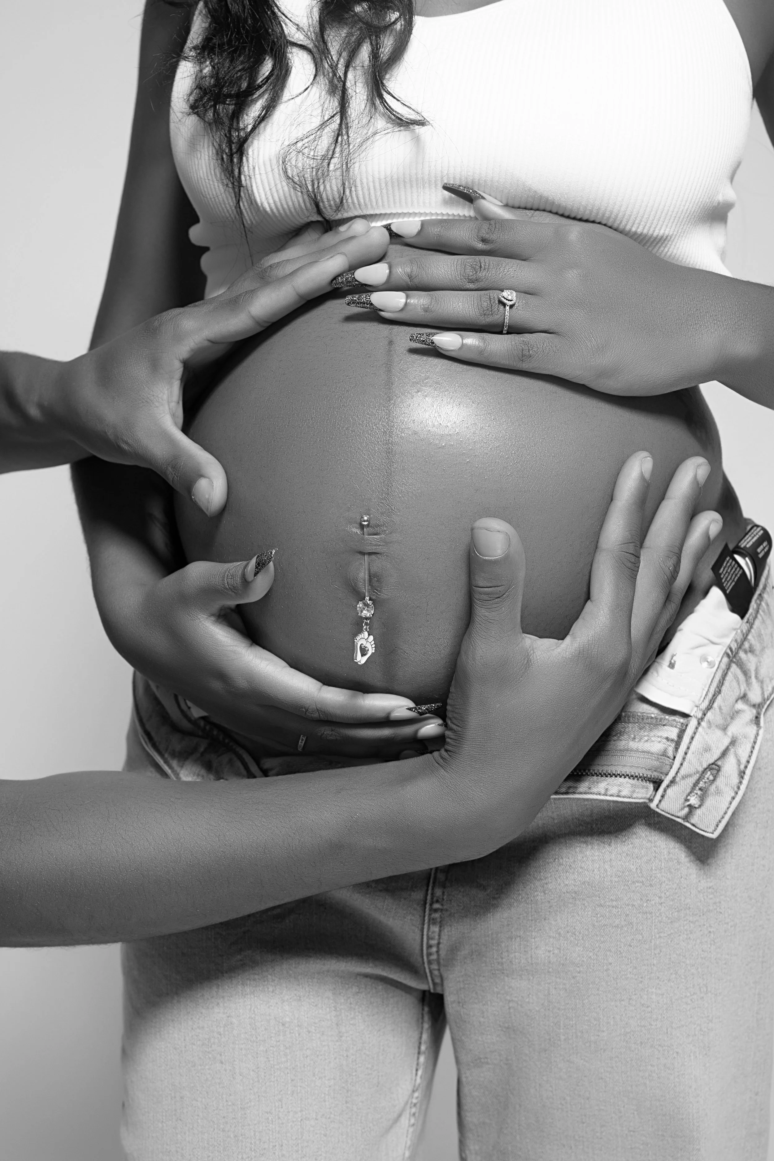 Close-up of multiple hands touching a pregnant woman's belly, some wearing rings, with the woman in a sleeveless top and jeans.
