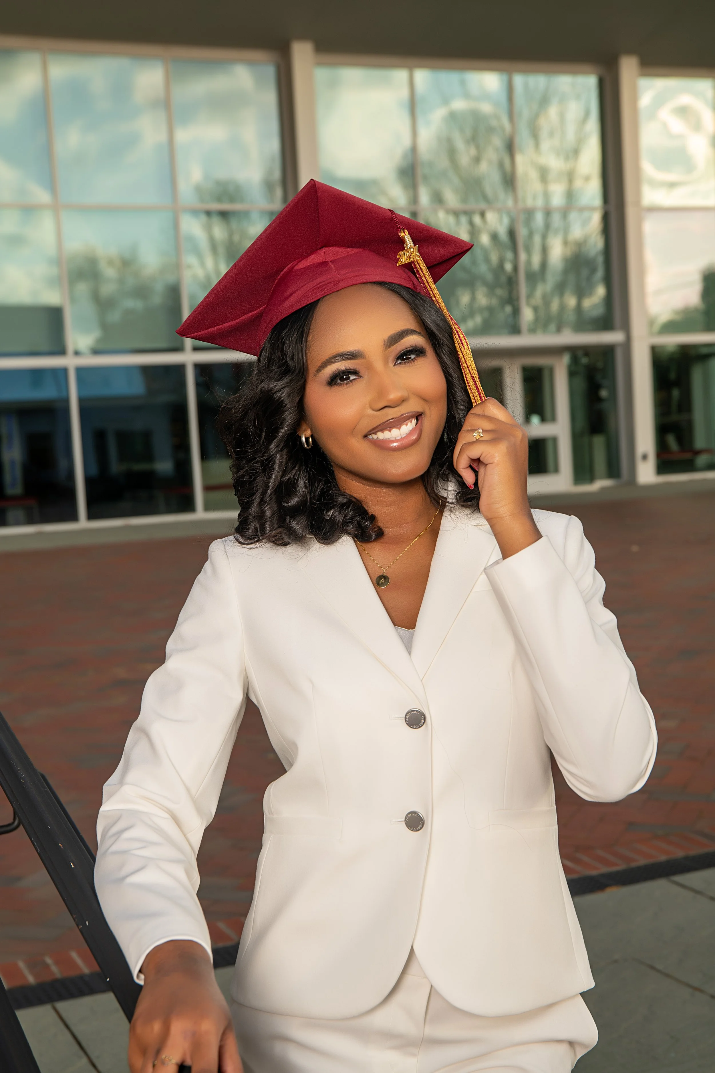 A woman in a white suit and red graduation cap is smiling outdoors, holding her cap with one hand.