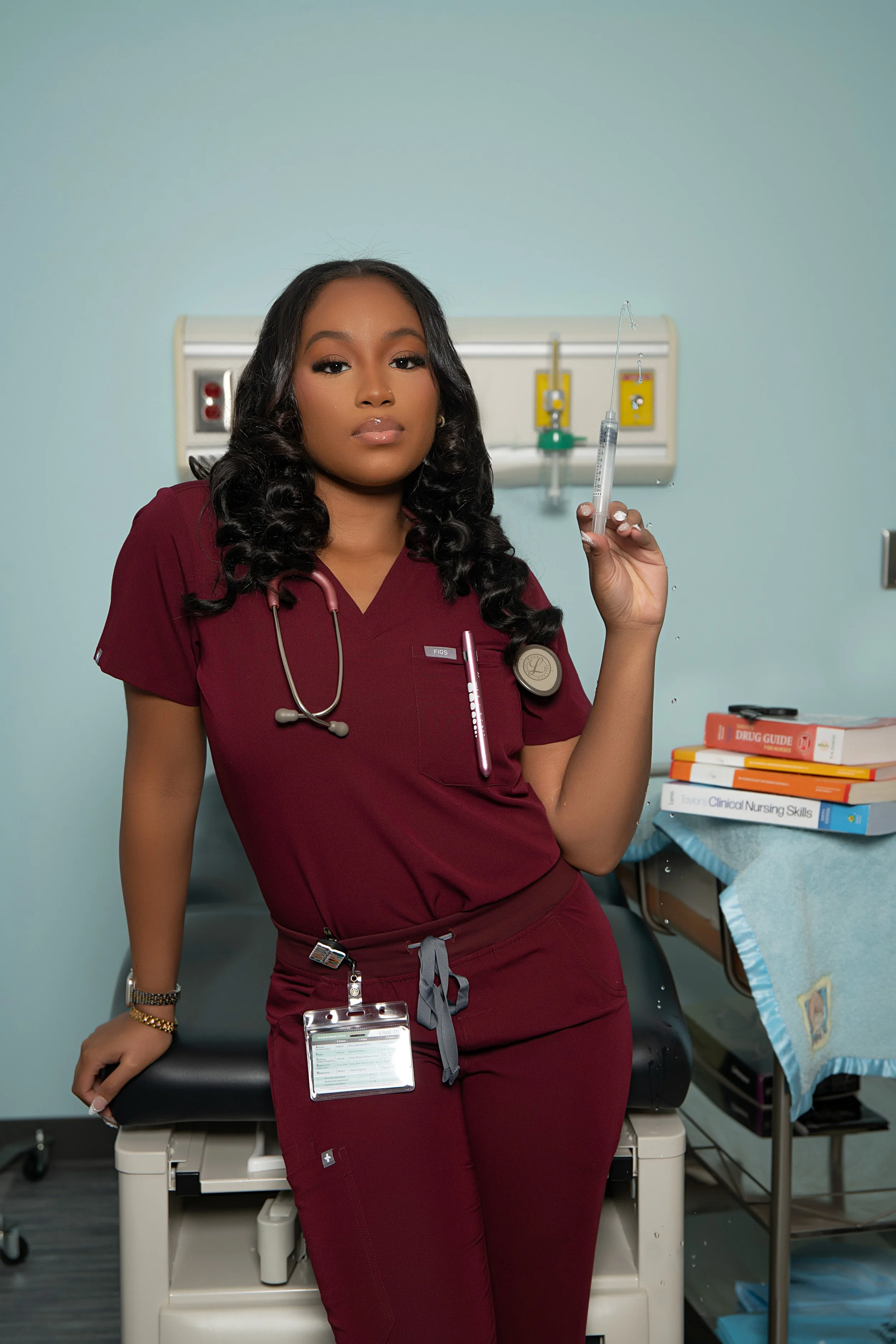 A female nurse with long dark hair in maroon scrubs, standing in a hospital room with medical equipment, holding a syringe, with a serious expression.