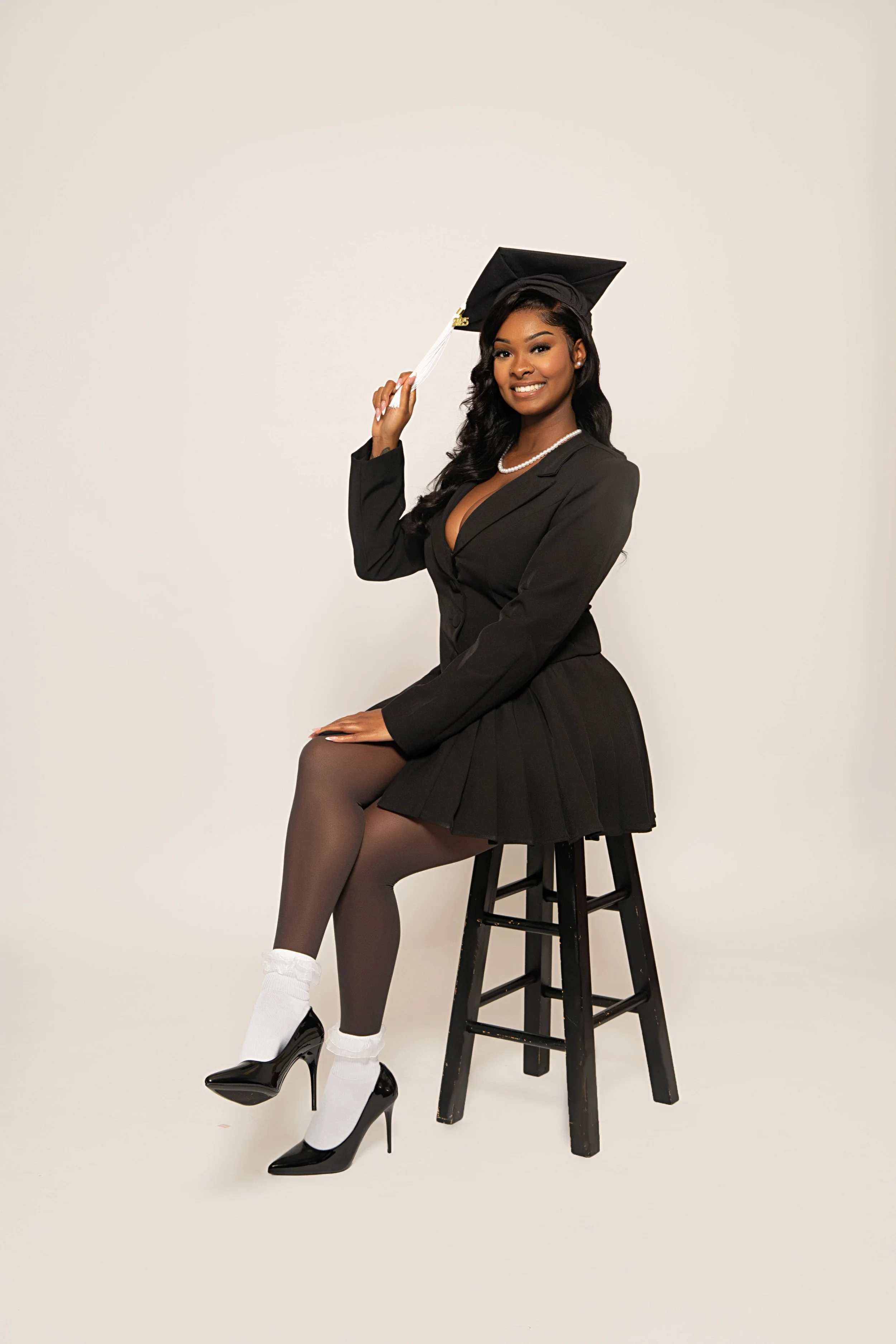 A young woman wearing a black graduation cap and gown, sitting on a black stool, smiling, with one hand adjusting her cap and the other resting on her lap. She is dressed in black stockings, white socks, black high heels, and is accessorized with a pearl necklace and earrings against a plain light background.
