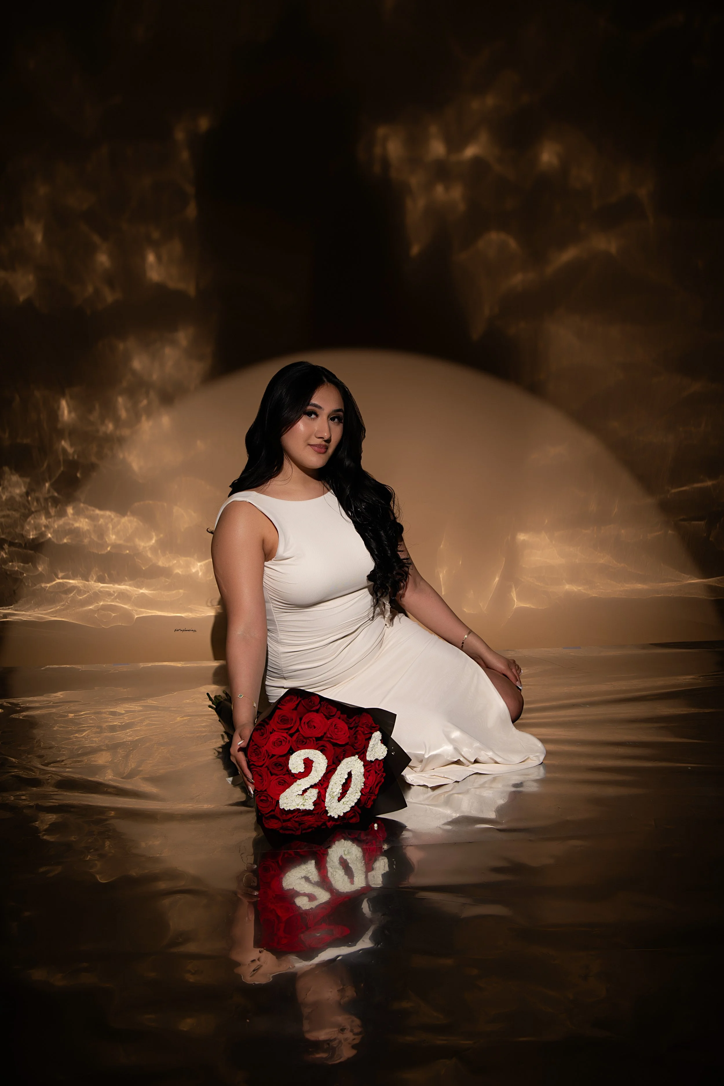 A young woman with long dark hair wearing a sleeveless white dress, sitting on a reflective surface in front of a dark, textured background. She holds a bouquet of red roses with a white '20' decoration on it, indicating a 20th celebration.
