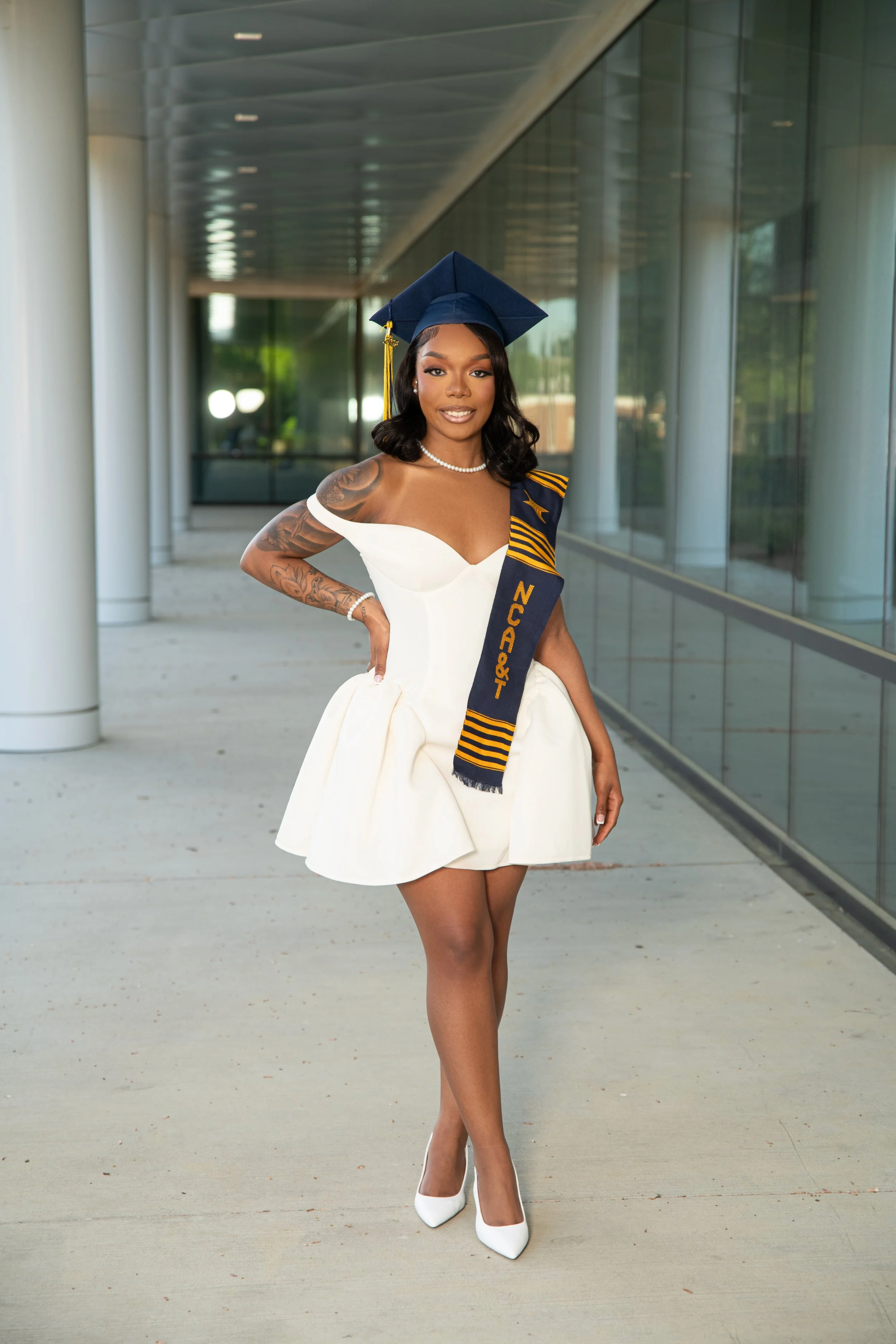 Woman wearing a white dress with shoulder cutouts, white high heels, a pearl necklace, and an American Graduation Cap with a sash that says 'NCA&T' standing outdoors in front of a modern building with glass windows.