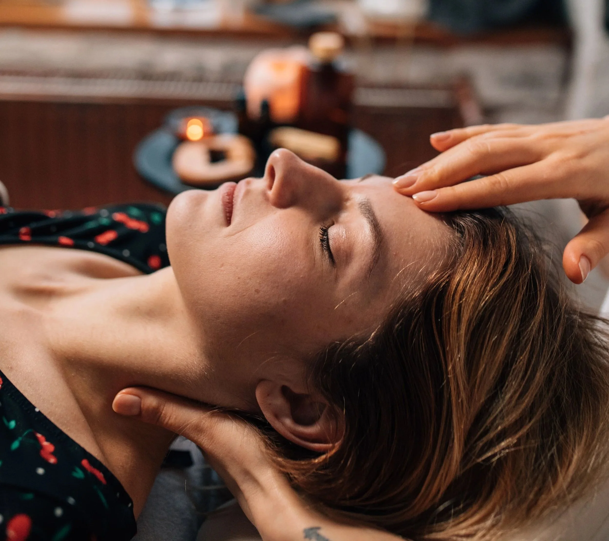 A person with short brown hair lying on her back with eyes closed, receiving a facial massage with fingers on her forehead, in a spa or wellness setting.