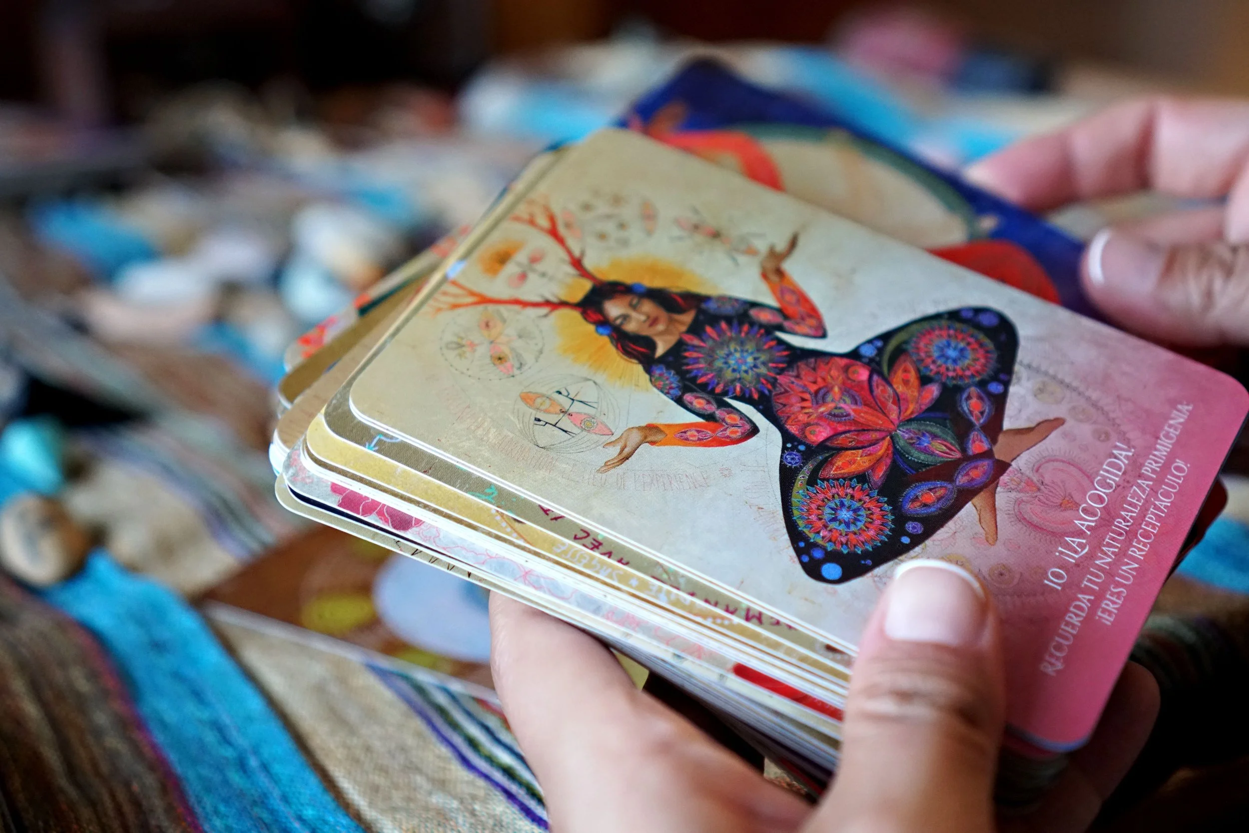 Close-up of a person's hand holding colorful tarot or oracle cards with intricate artwork, featuring a woman with butterfly wings and a crown, with blurred objects in the background.