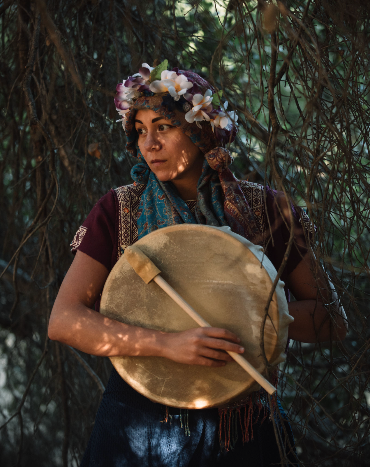 A woman holding a round drum with a stick while standing among tree branches, wearing a floral headpiece and traditional clothing.