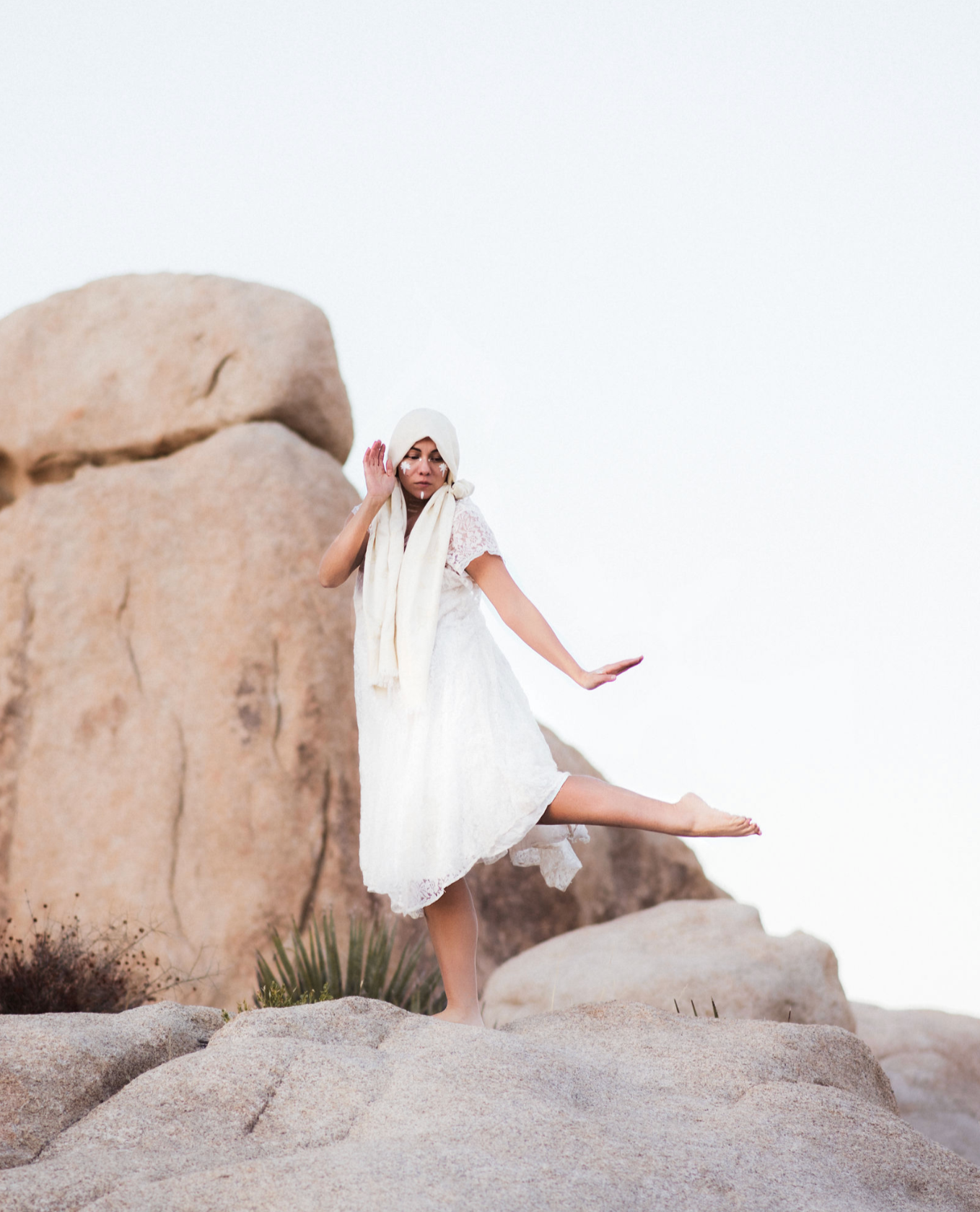 A woman in a white dress with a hoodie is sitting on rocks and balancing on one leg while extending her other leg out, with her arms outstretched. She is outdoors among large rocks and desert plants, and the sky is overcast.