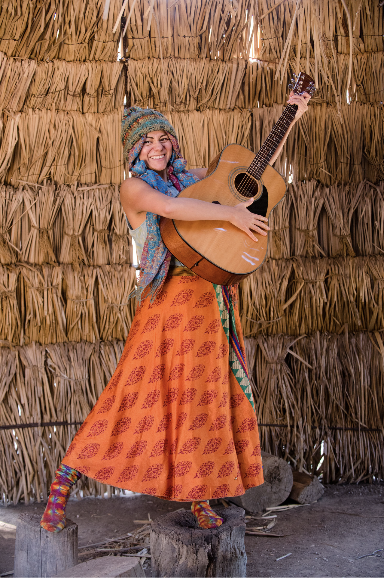 A woman wearing a colorful hat and a long orange skirt, holding an acoustic guitar, standing on wooden stumps inside a structure made of bundled straw or reeds.