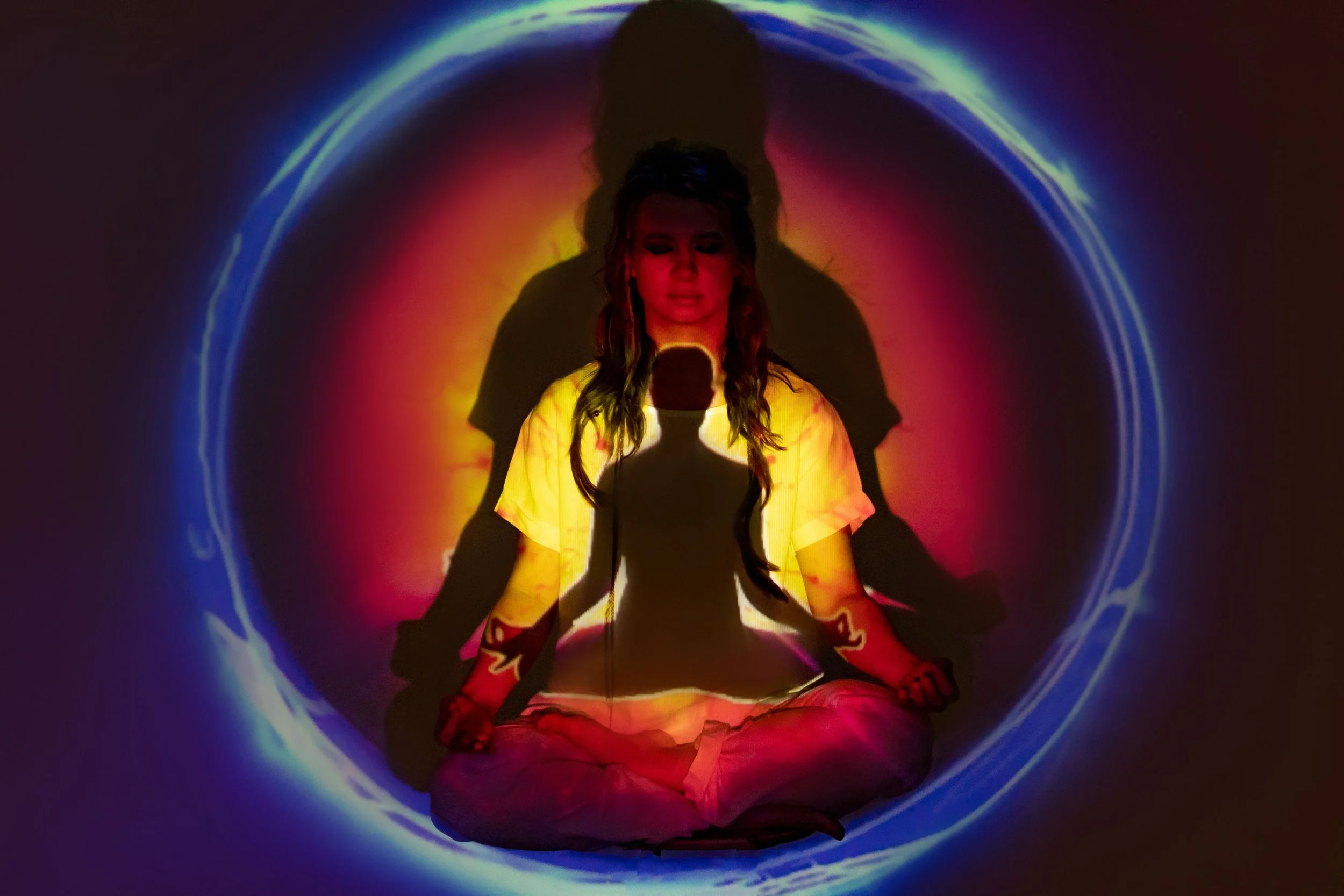 A woman sitting cross-legged in the dark, with colorful projection lights creating a circular glow around her, casting her outline and a shadow on the wall behind.