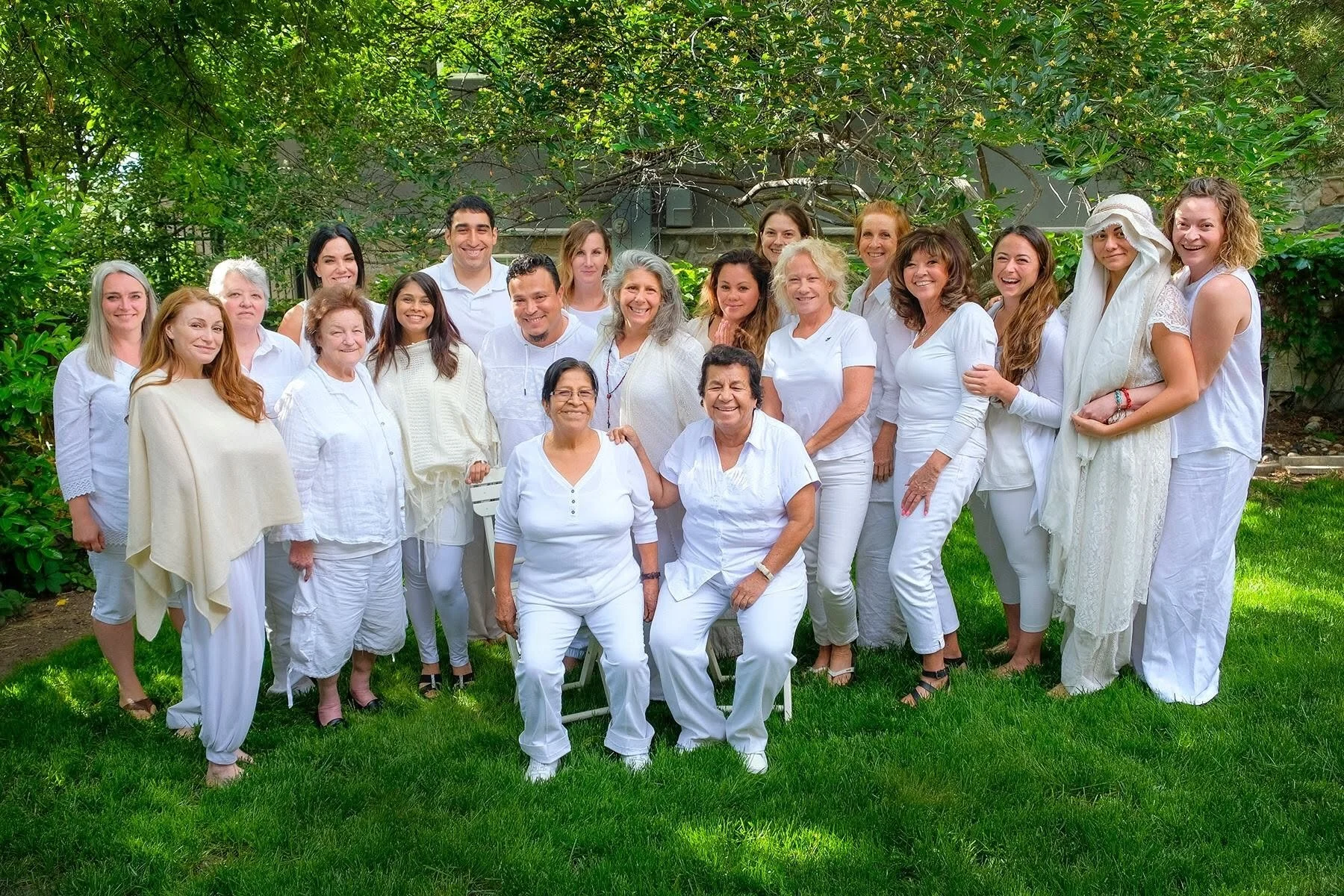 Group of people dressed in white, standing outdoors on green grass, with lush trees and foliage in the background, smiling at the camera.