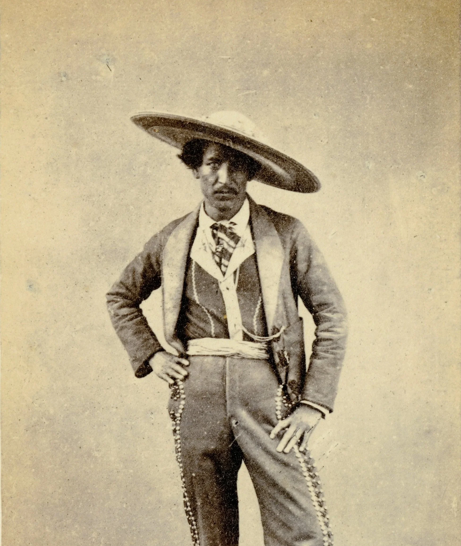 A black and white historical photograph of a man wearing a large, wide-brimmed hat, a suit with a scarf around his neck, and holding a string of beads.