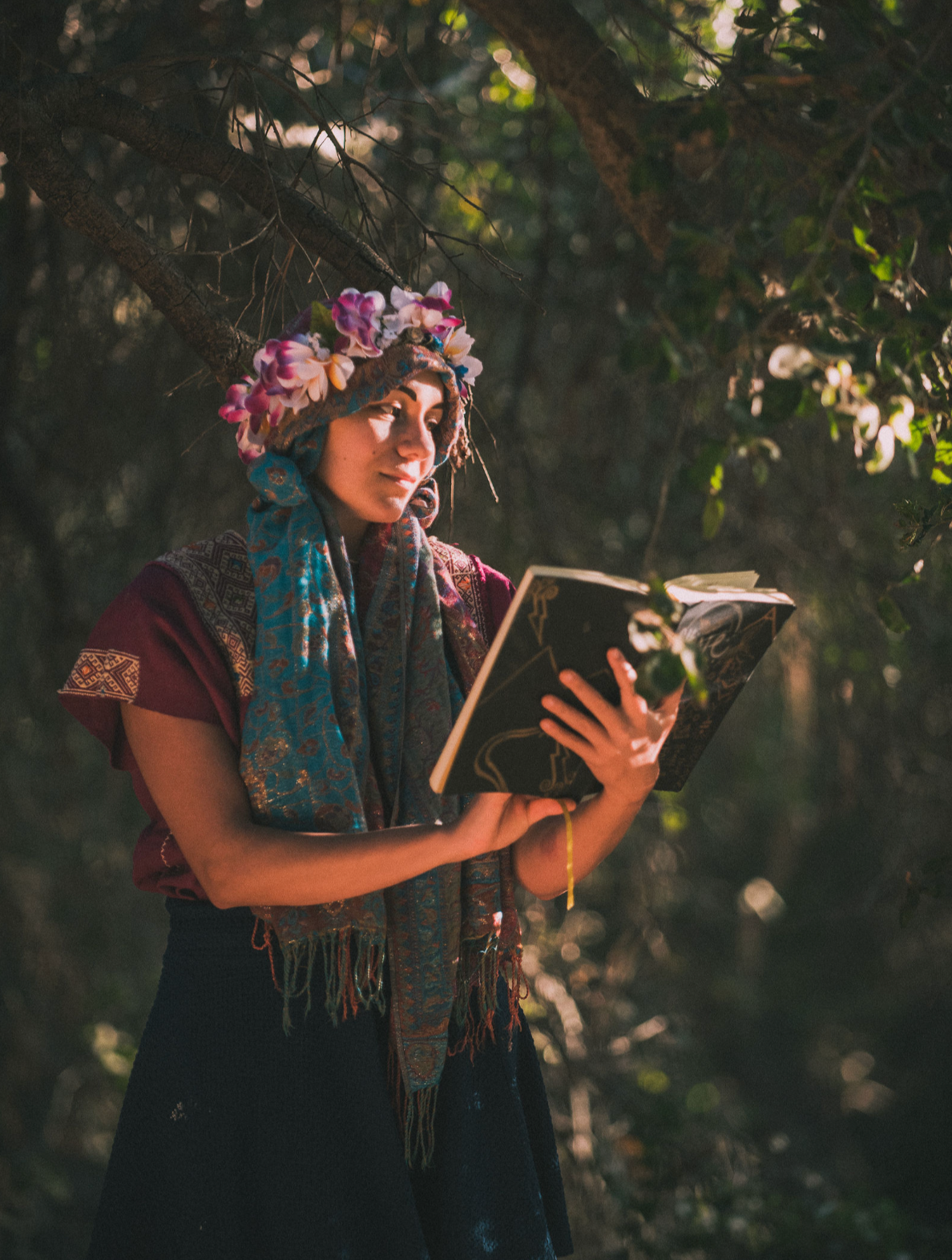 A woman dressed in traditional clothing with a floral headdress, holding and reading a book outdoors amid trees and natural light.