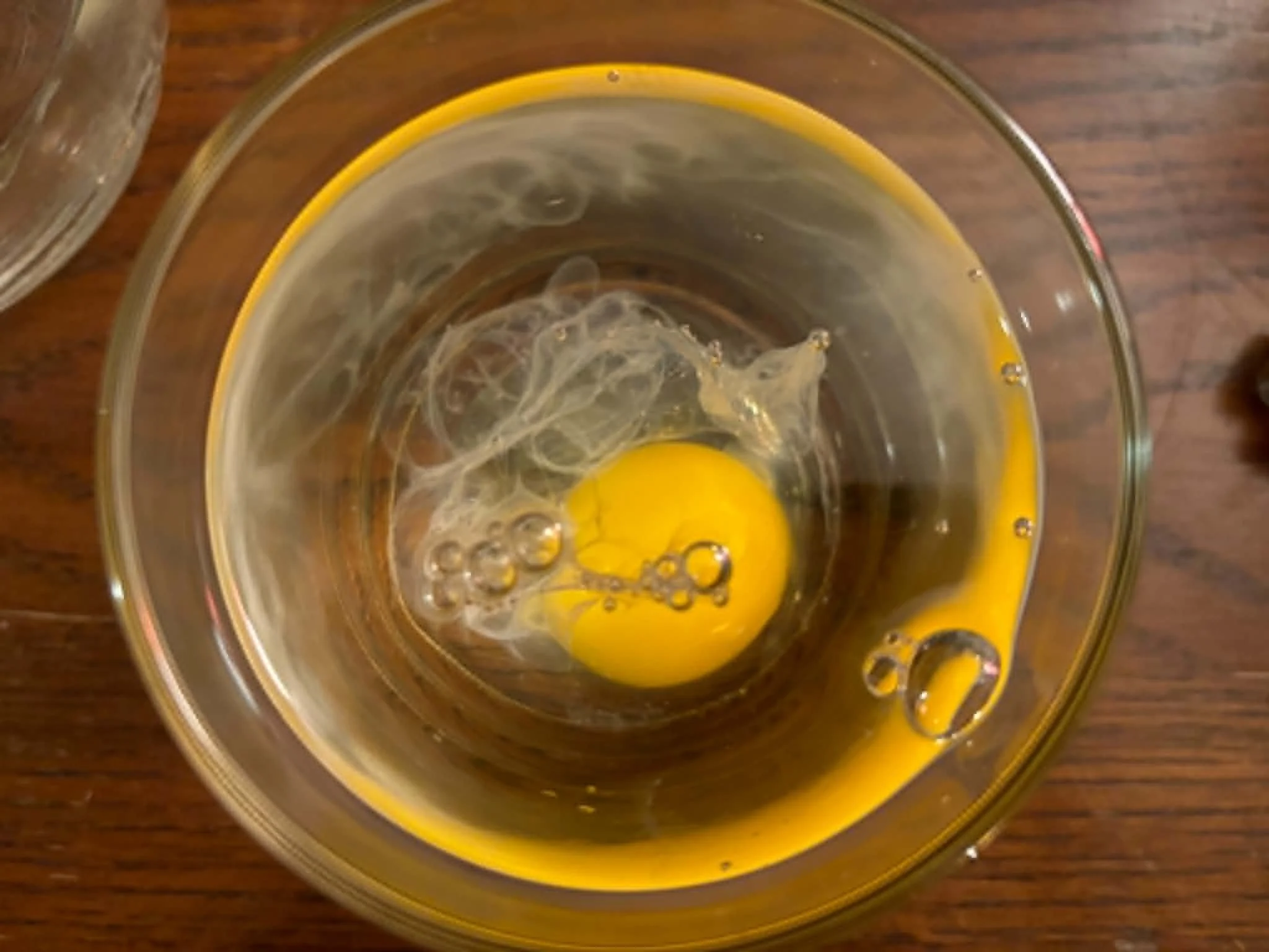 A raw egg cracked into a clear glass measuring cup, with the yolk visible at the bottom, on a wooden surface.