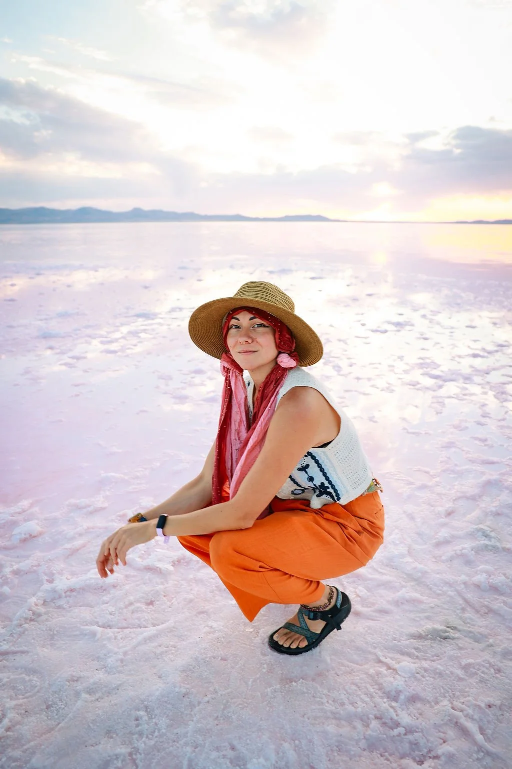Woman squatting on pink salt flats wearing a wide-brimmed straw hat, orange pants, sleeveless top, and sandals, with a scenic sunset sky in the background.