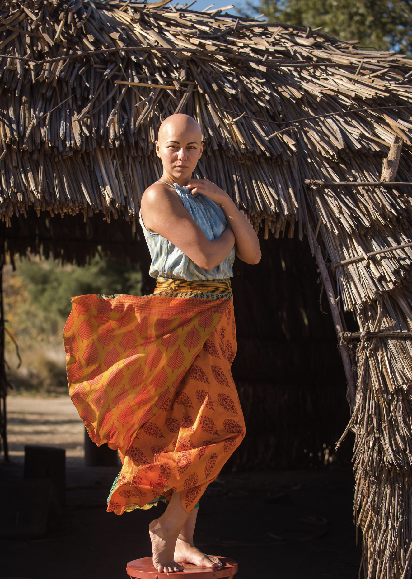 A woman with alopecia standing barefoot on a red stool in front of a thatched hut, wearing a blue top and orange traditional skirt with yellow and red pattern, outdoors on a sunny day.