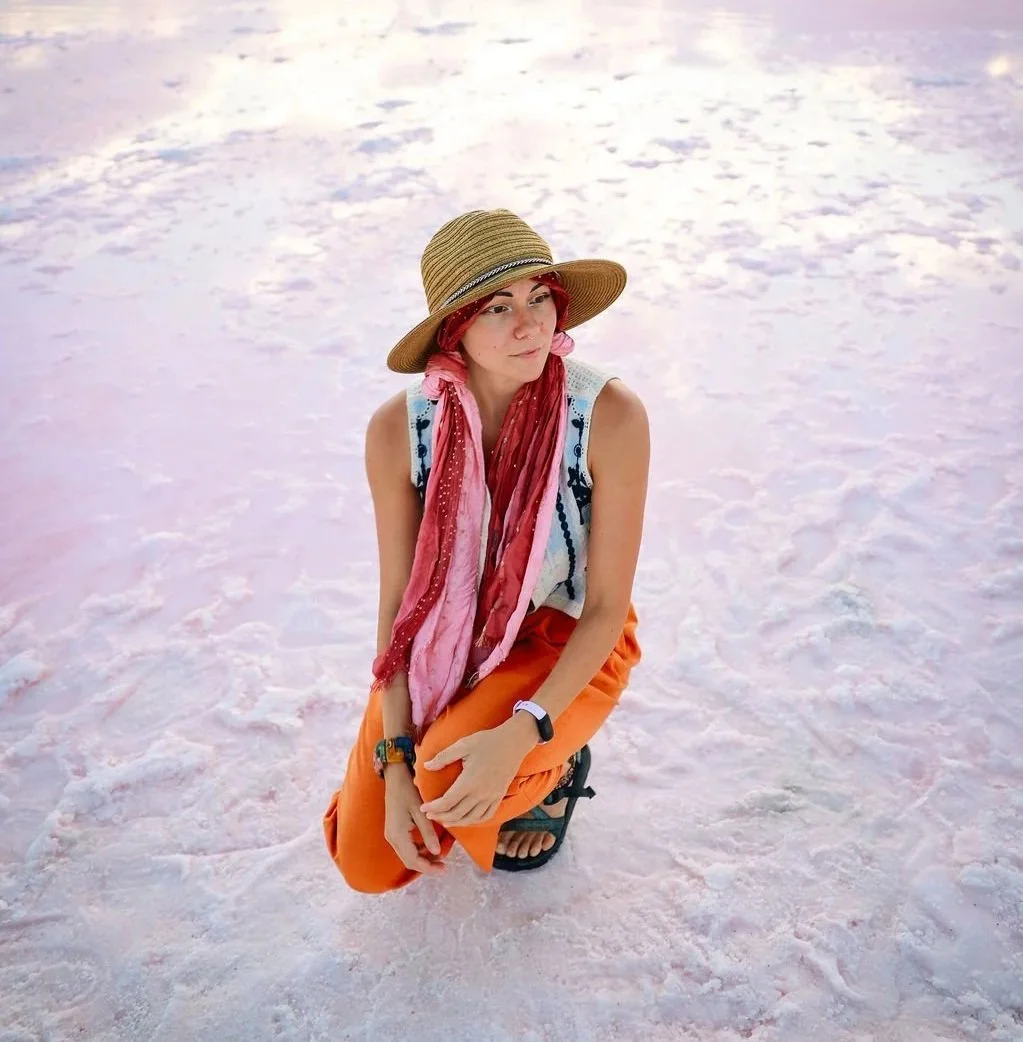 A woman with red hair, sitting on the salt flats, wearing a wide-brimmed straw hat, orange pants, a sleeveless top, a pink scarf, and sandals.