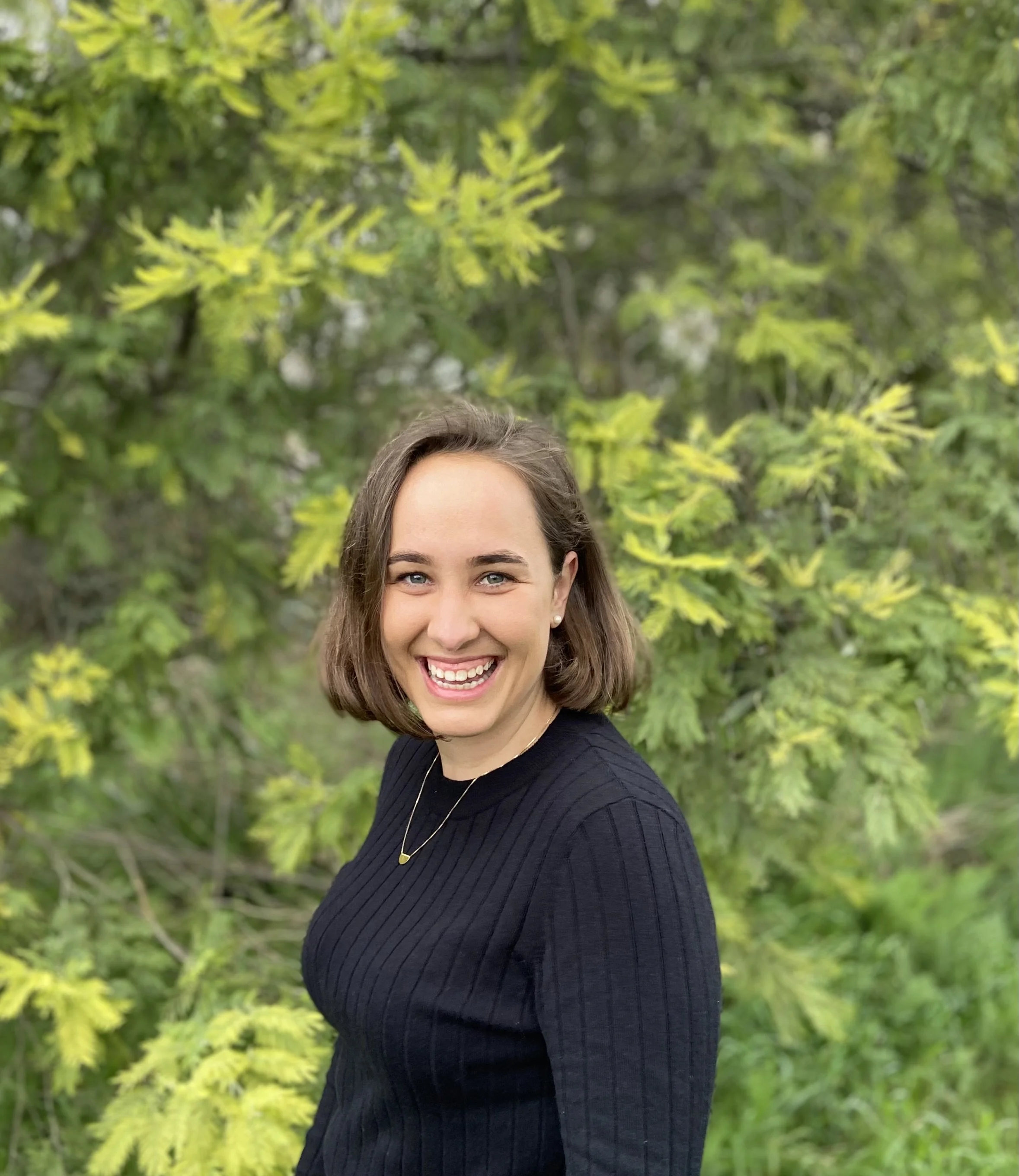 Close-up of Kate Boersen smiling wearing a black top, standing outside with green trees in the background.