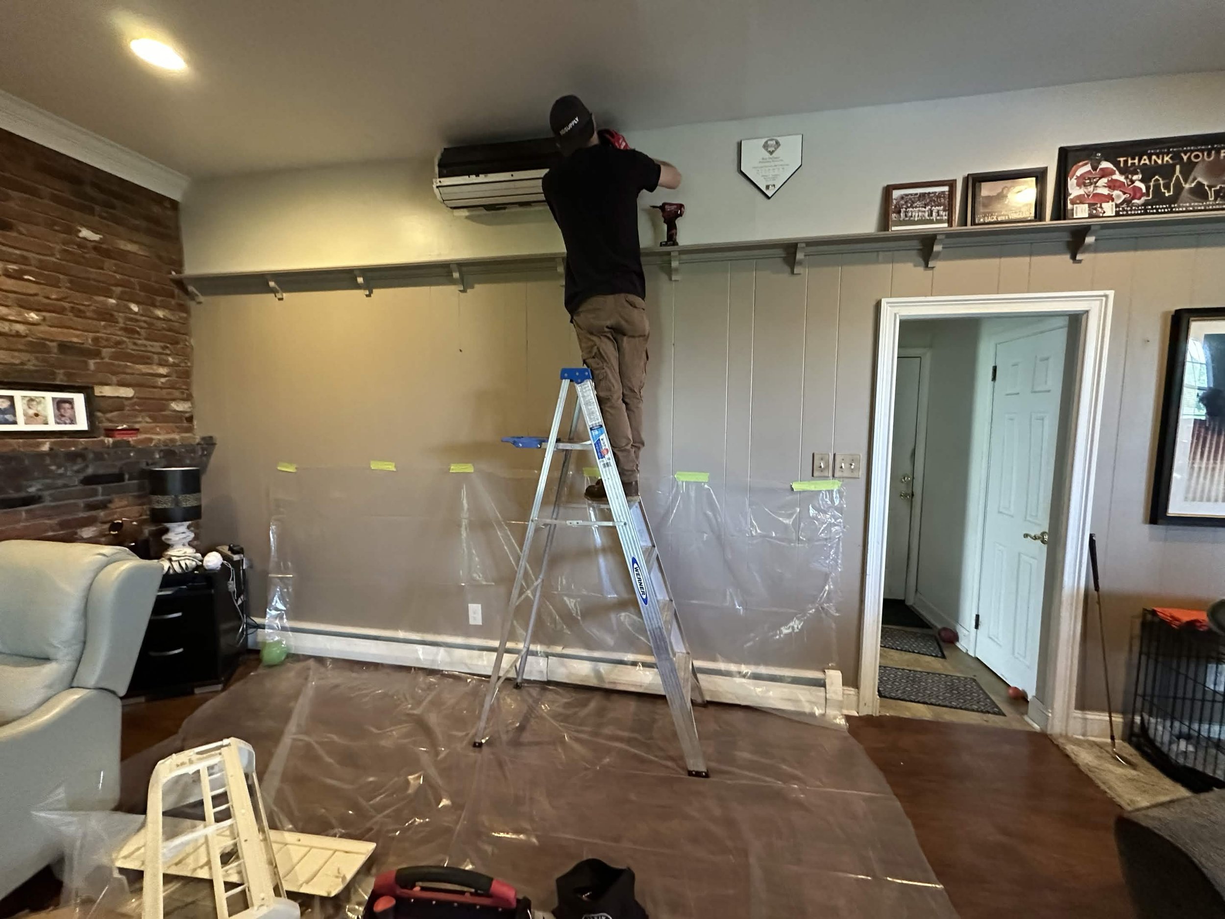 A person standing on a step ladder installing or repairing an air conditioning unit in a living room, with plastic sheeting on the floor and various tools nearby.