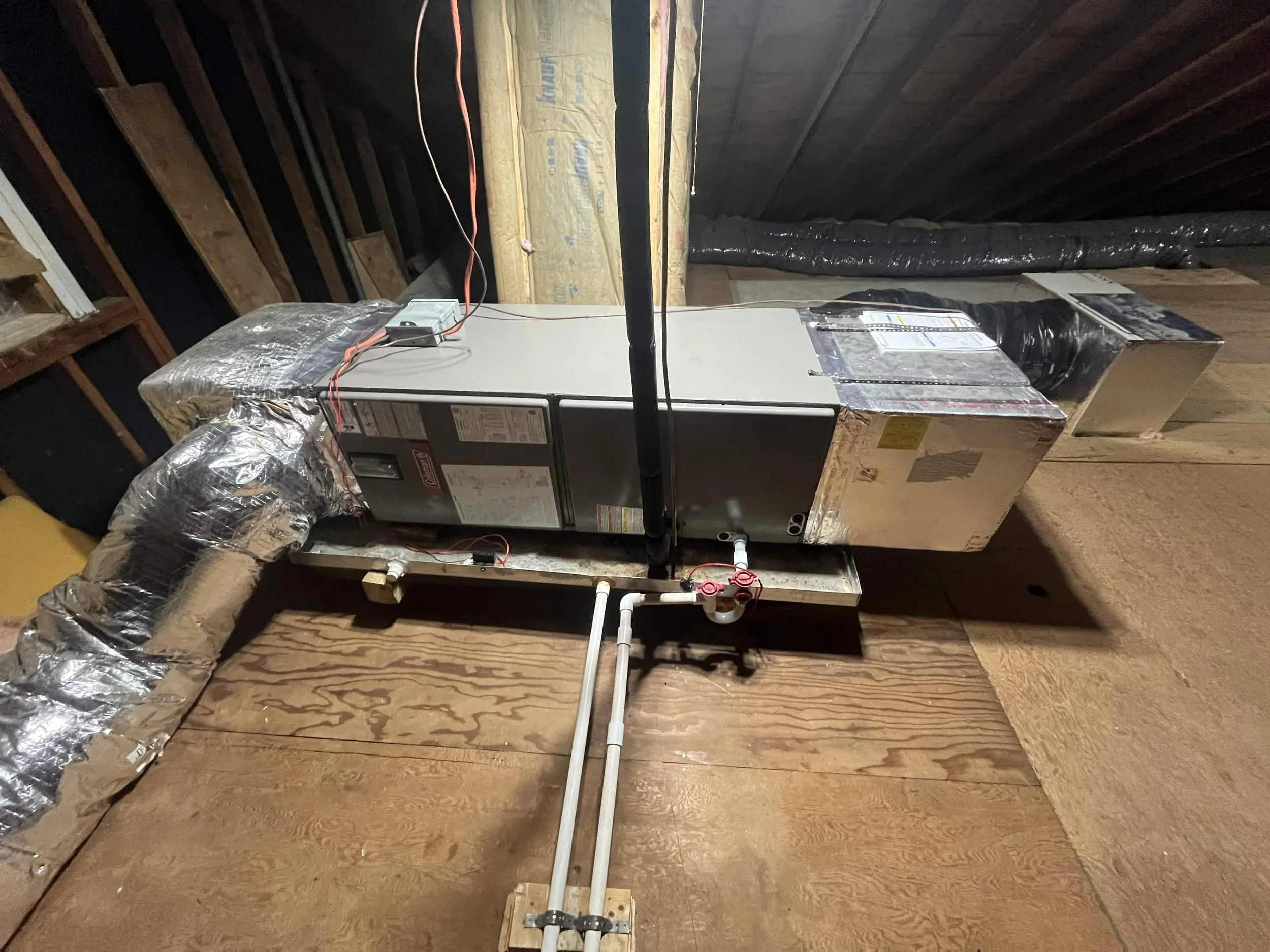 A furnace or HVAC system set up in an unfinished attic space with exposed wooden beams and insulation on the walls.