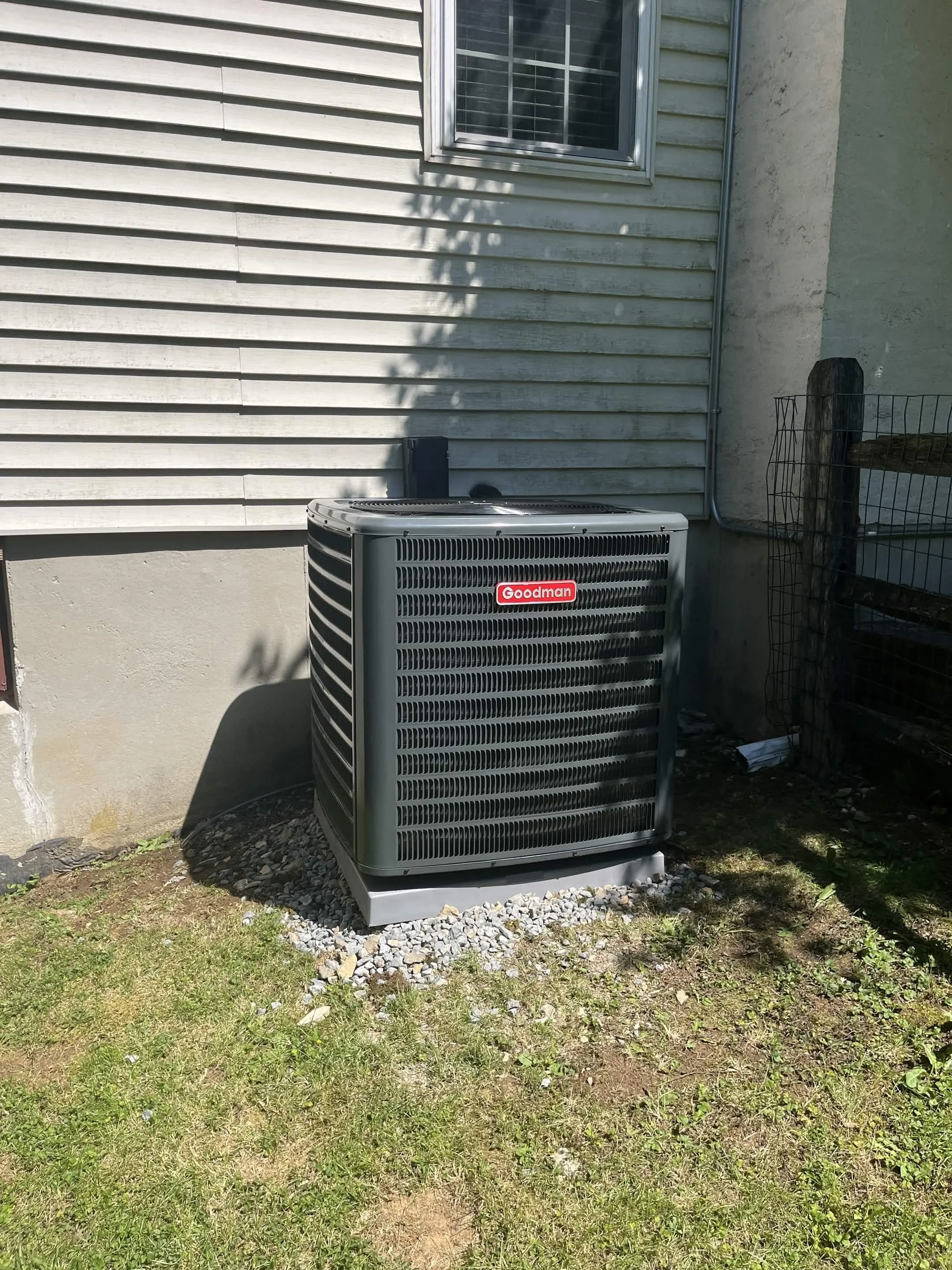 An outdoor air conditioning unit labeled 'Goodman' installed next to a house with vinyl siding, a window, and a wire fence.