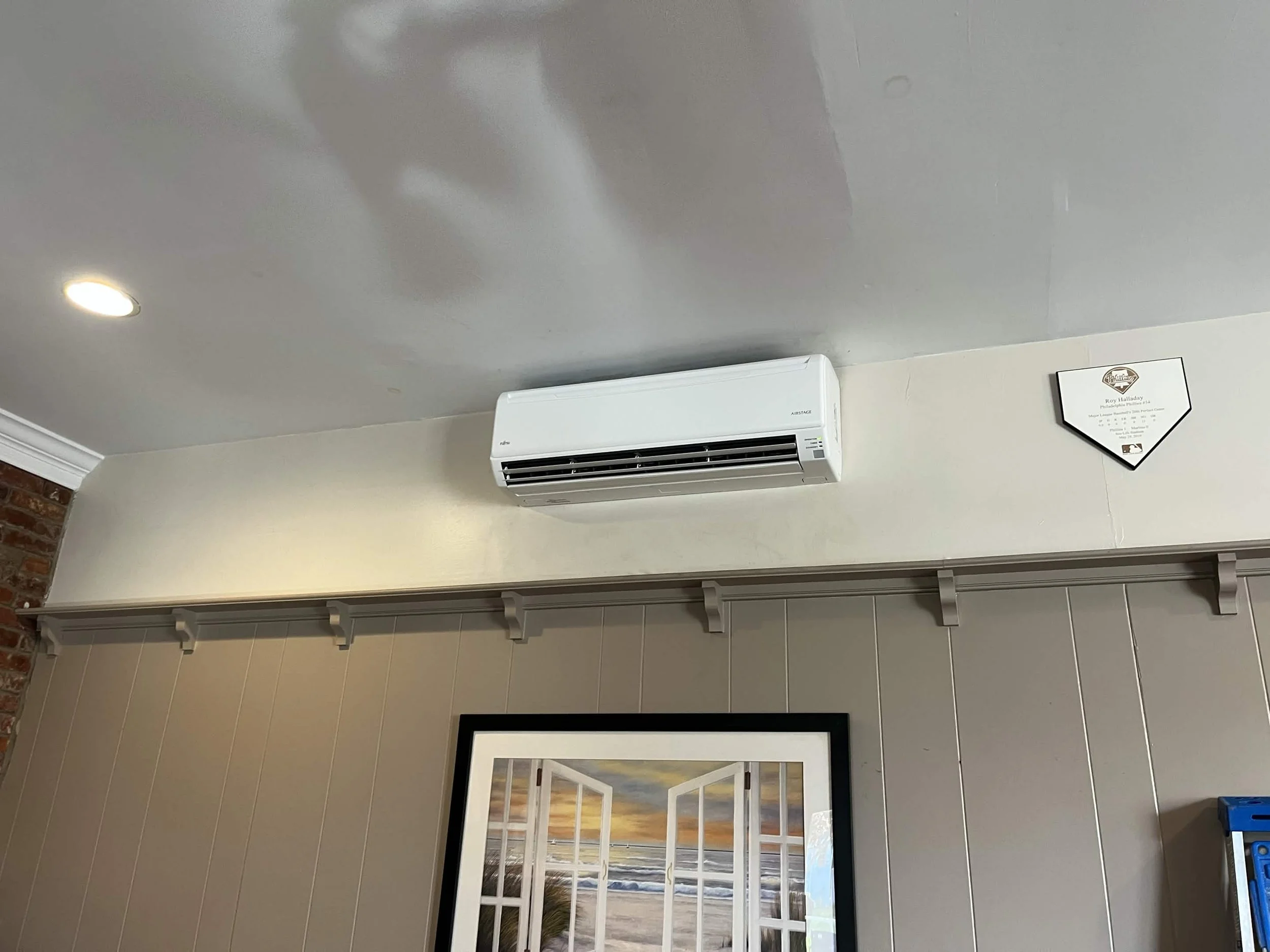 A room with a white wall-mounted air conditioner, framed artwork, and a plaque on the wall. The ceiling has a recessed light, and part of a brick wall is visible on the left.