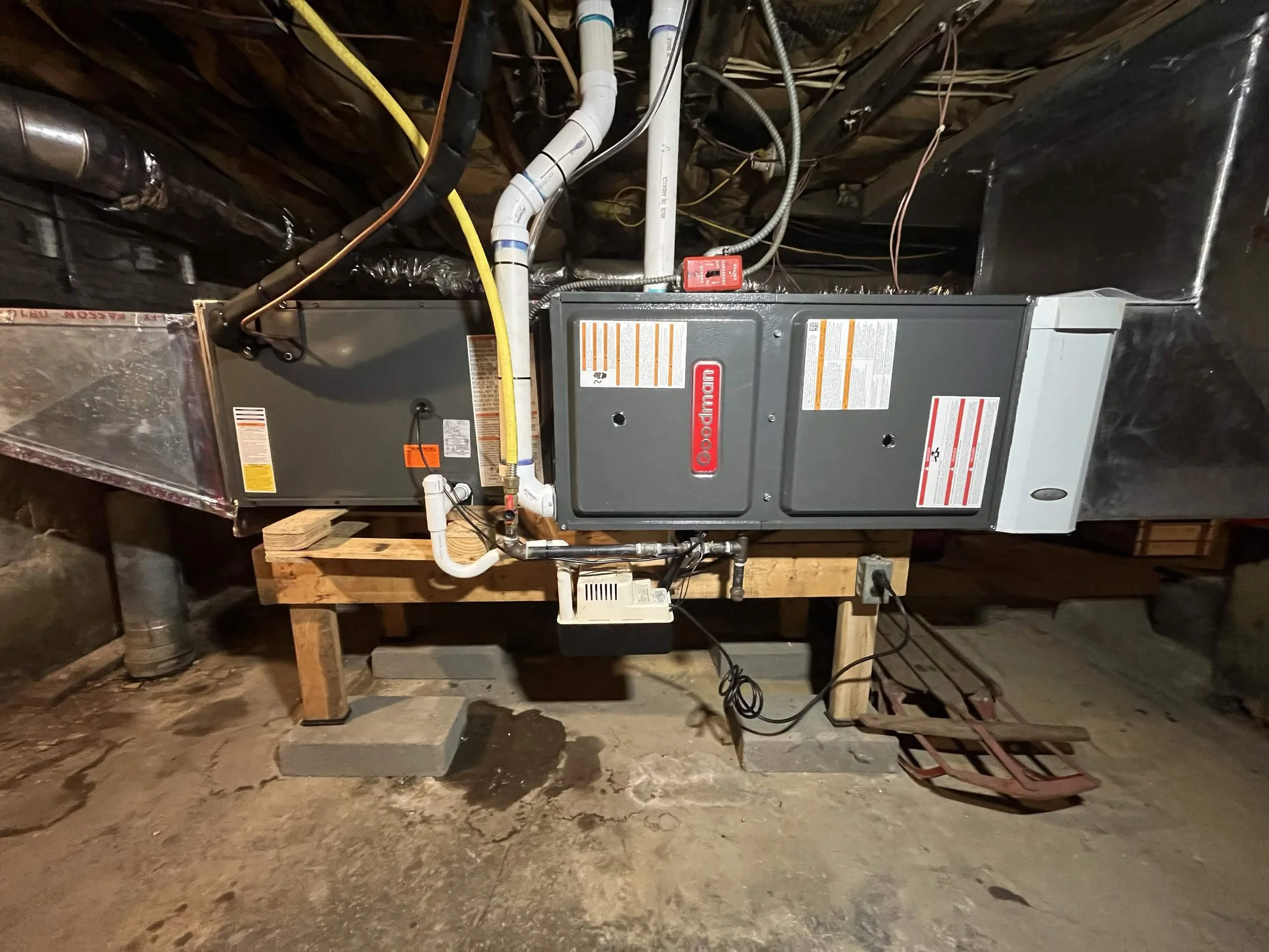 A central heating furnace installed on a concrete floor in a basement, with a wooden platform supporting it and various pipes and wires connected to it.