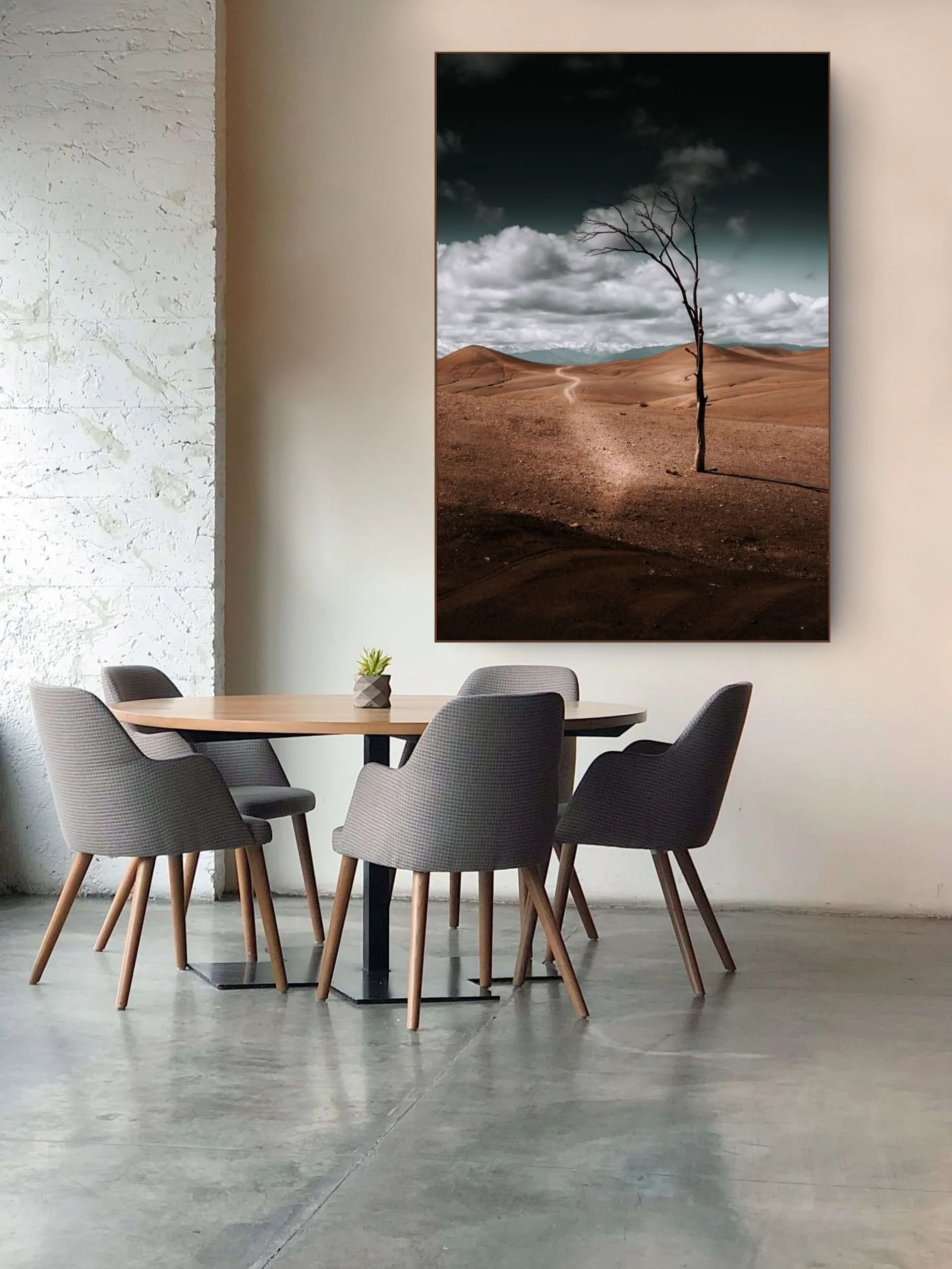 Modern conference room with a round table, six gray fabric chairs with wooden legs, a small potted plant, and a large landscape poster of a barren desert scene with a lone leafless tree on a wall.