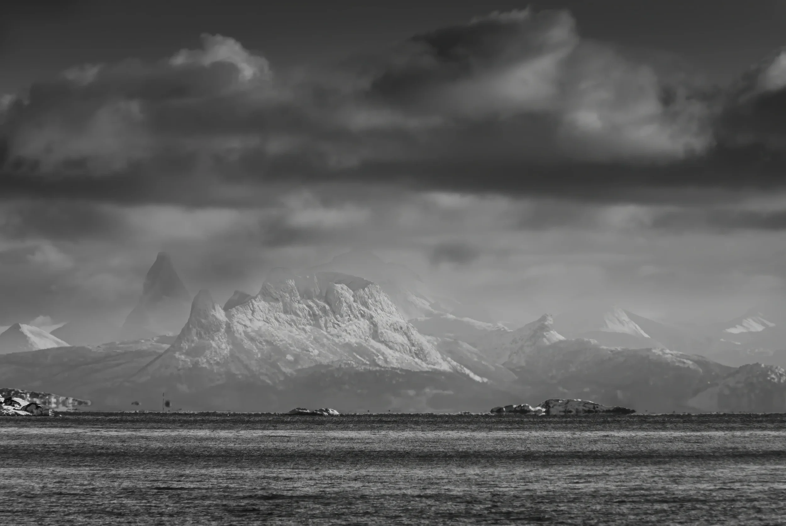 Jagged snow-covered peaks above a Norwegian fjord under a dramatic stormy sky, black and white photography.