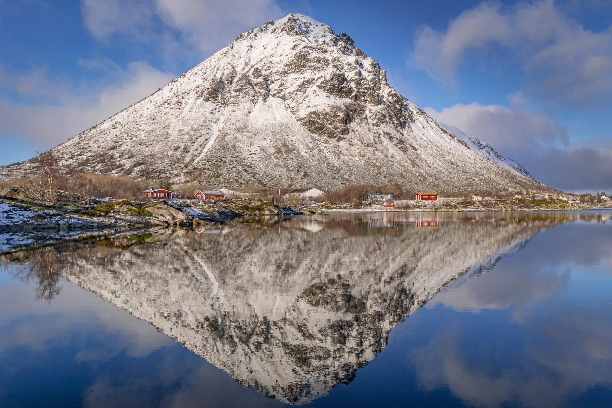 Snow-covered mountain with a reflection in a body of water below, small houses at the base of the mountain, cloudy blue sky.