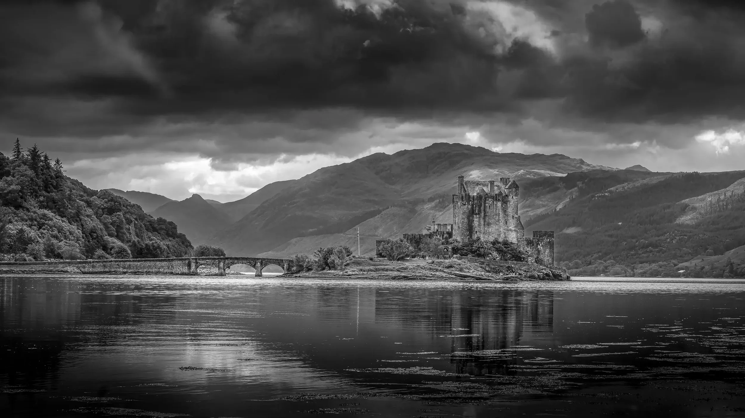 Eilean Donan Castle in Scotland reflected in still water under a dramatic black and white sky.