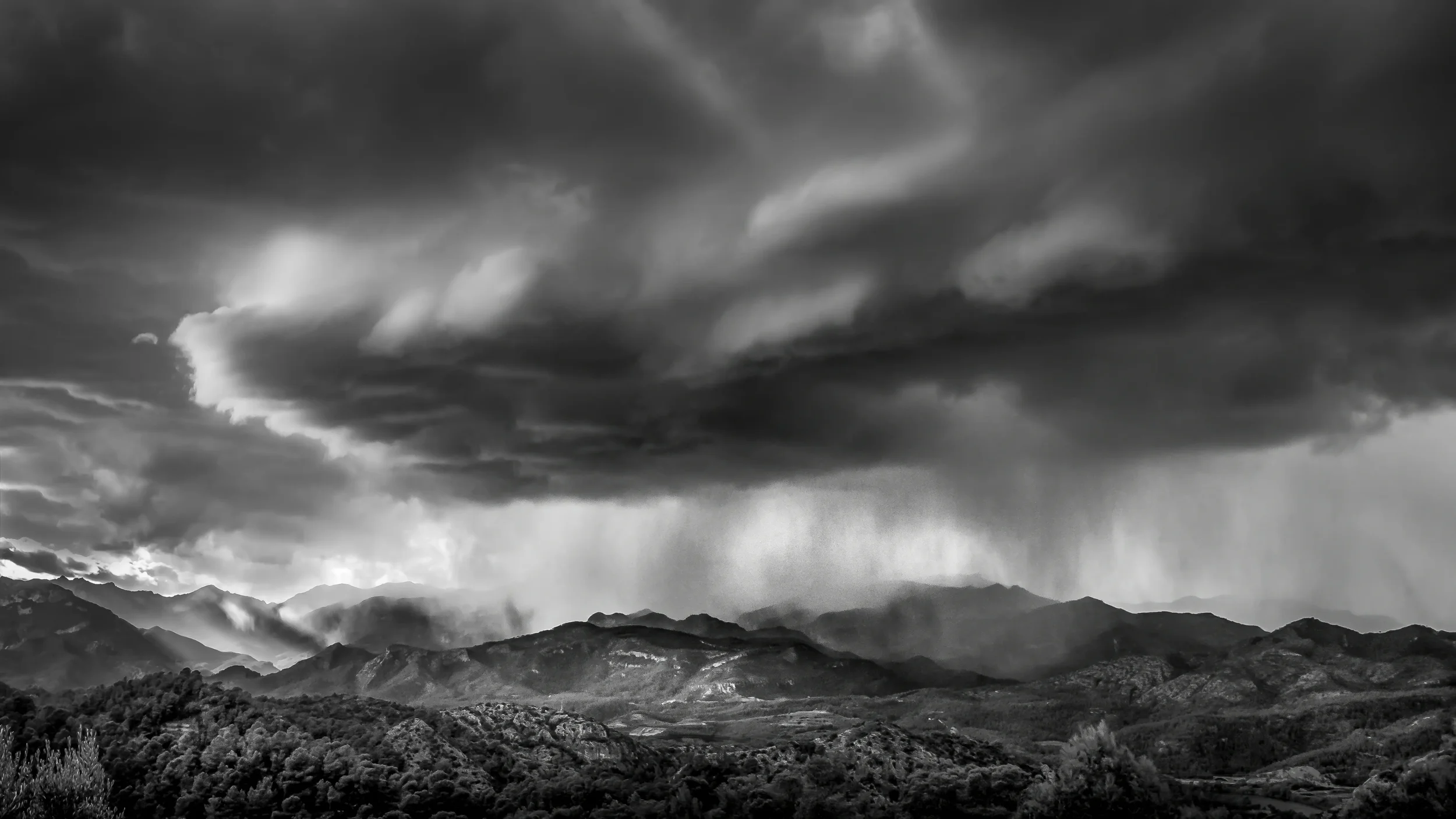 Dramatic storm clouds with visible rainfall over a dark mountain range, black and white photography.