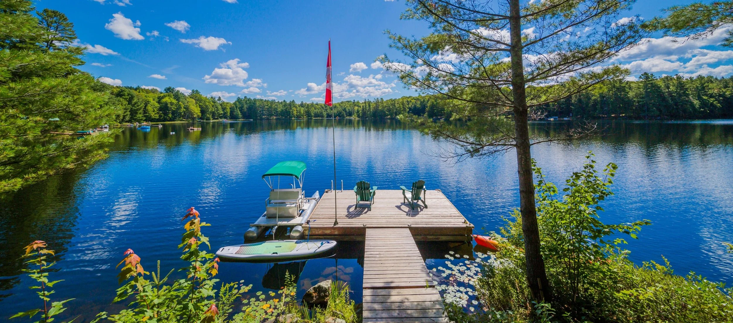 A peaceful lakeside scene with a wooden dock extending into the water, two green Adirondack chairs, a small boat with a green canopy, and a red flag on a pole. Surrounding trees with green leaves frame the bright blue sky with scattered clouds.