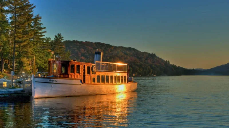 SS Bigwin on a lake at sunset, with tree-lined hills in the background and the water reflecting the sunlight.