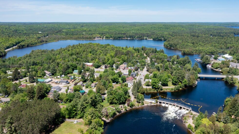 Aerial view of Baysville in Lake of Bays, ON surrounded by dense forests, with multiple lakes and rivers, and a dam releasing water in the foreground.