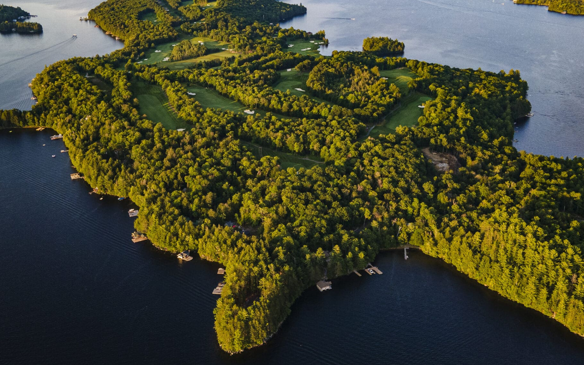 Aerial view of Bigwin island in Lake of Bays, ON with a golf course, surrounded by water with several boats docked around its edges.