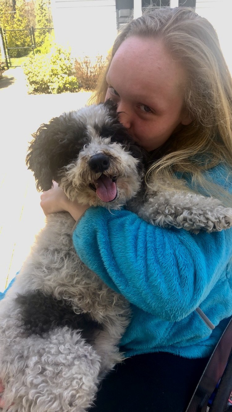 A woman with long blonde hair hugging a fluffy black and white dog outdoors.