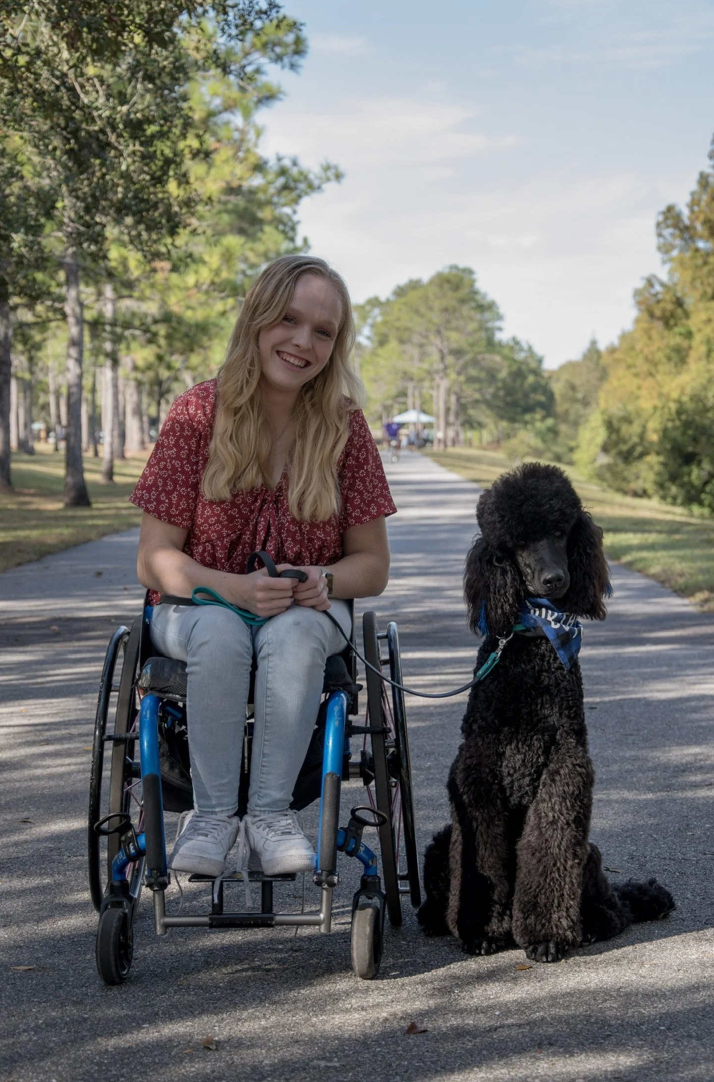 A young woman with long blonde hair sitting in a wheelchair, smiling, holding a leash attached to a large black and gray poodle sitting beside her on a paved walking path in a park with tall trees and a blue sky in the background.
