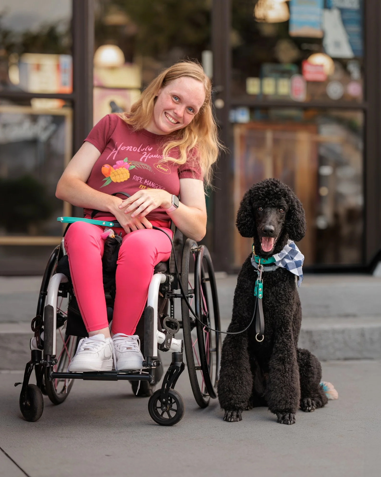 A young woman with red hair in a wheelchair next to a large black poodle dog. The woman is smiling and wearing a red t-shirt with tropical fruit and flower graphics and pink pants, outdoors in front of a building with large glass windows.