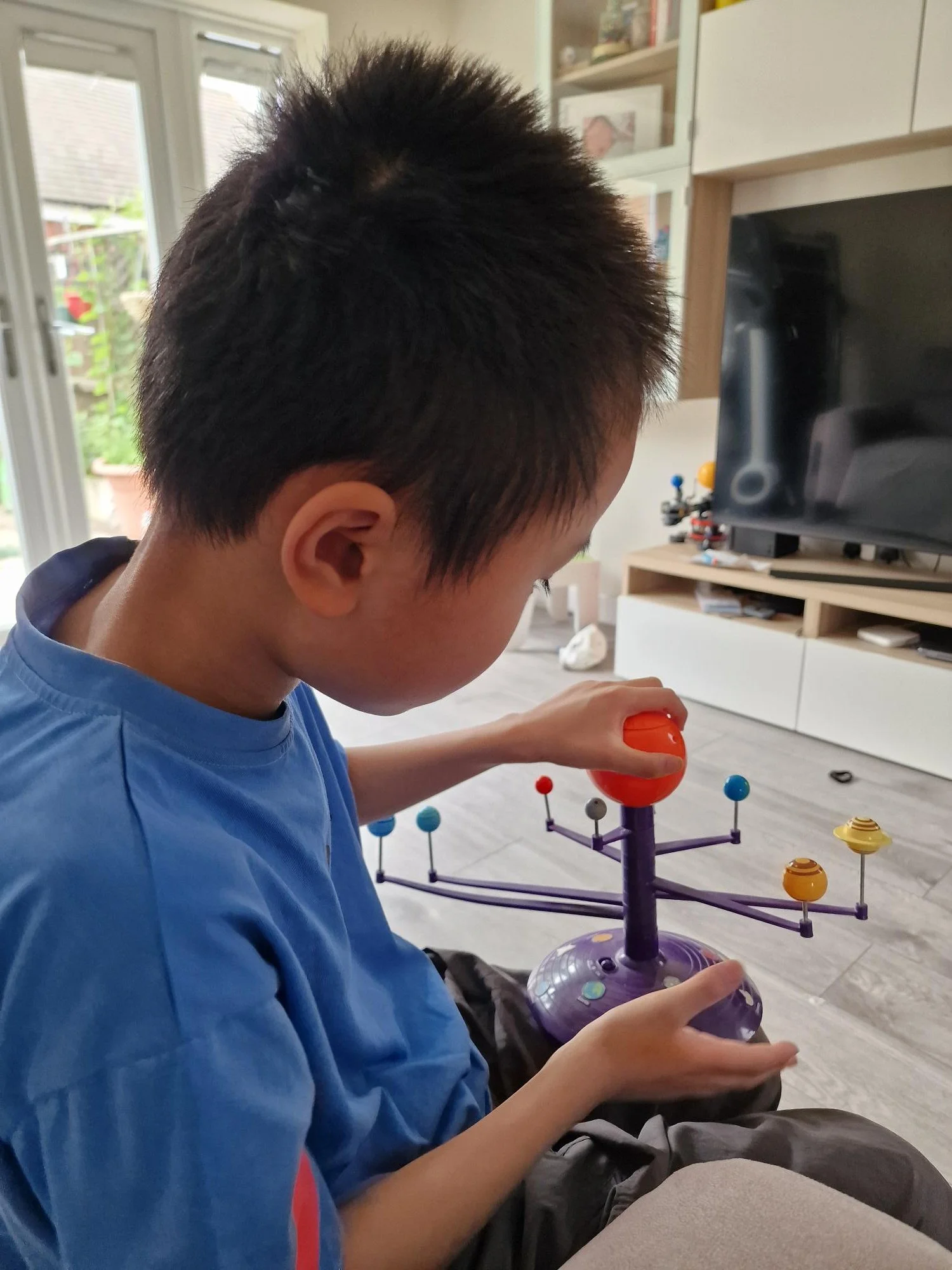 A child with short dark hair playing with a sci-fi themed board game inside a living room. The game features a purple base and colorful orbital pieces. Behind, a TV and shelves with books and toys are visible.